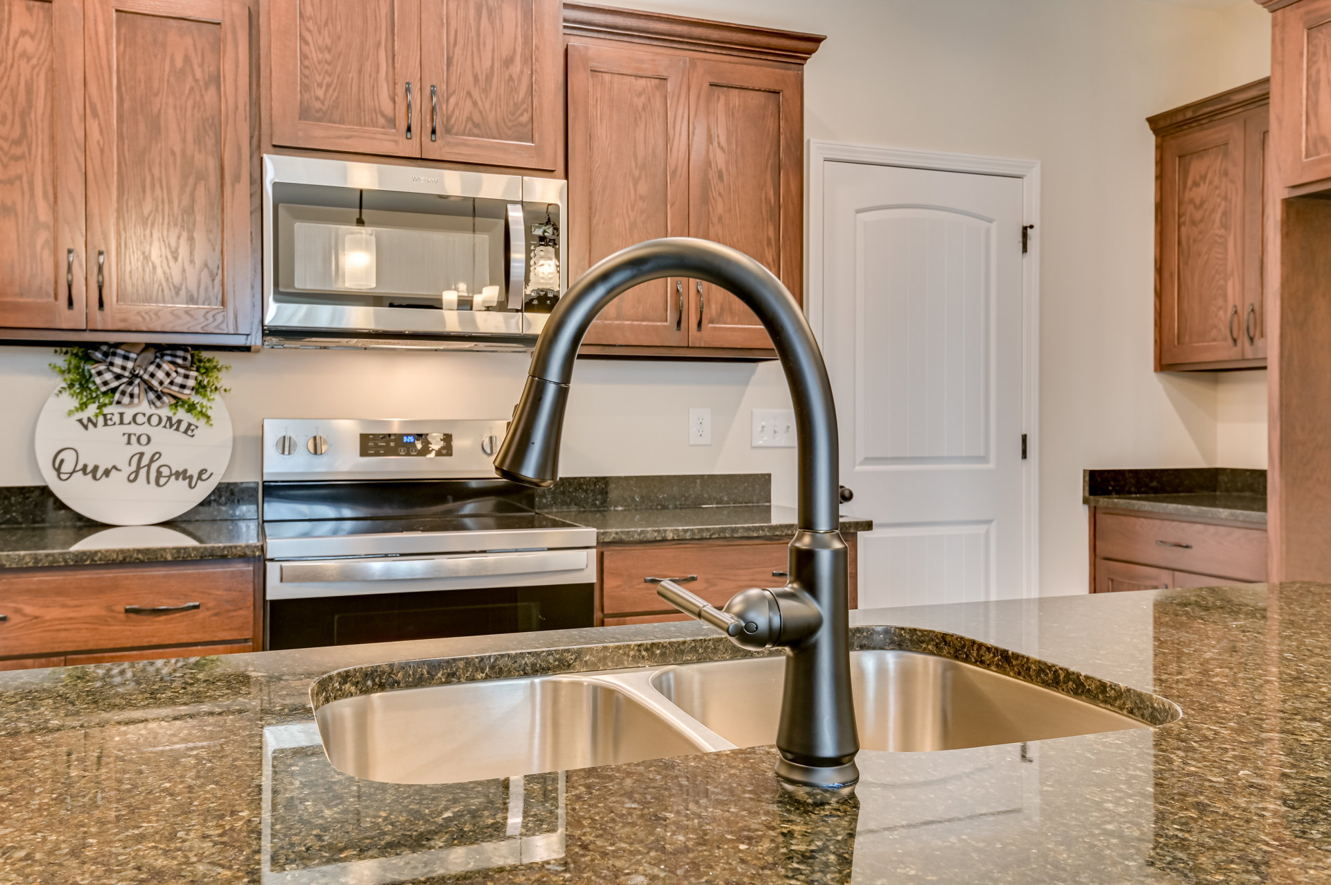 Modern kitchen featuring a stainless steel sink with black faucet, white cabinetry, stone countertop, built-in oven, and microwave; candles and decorative sign displayed near