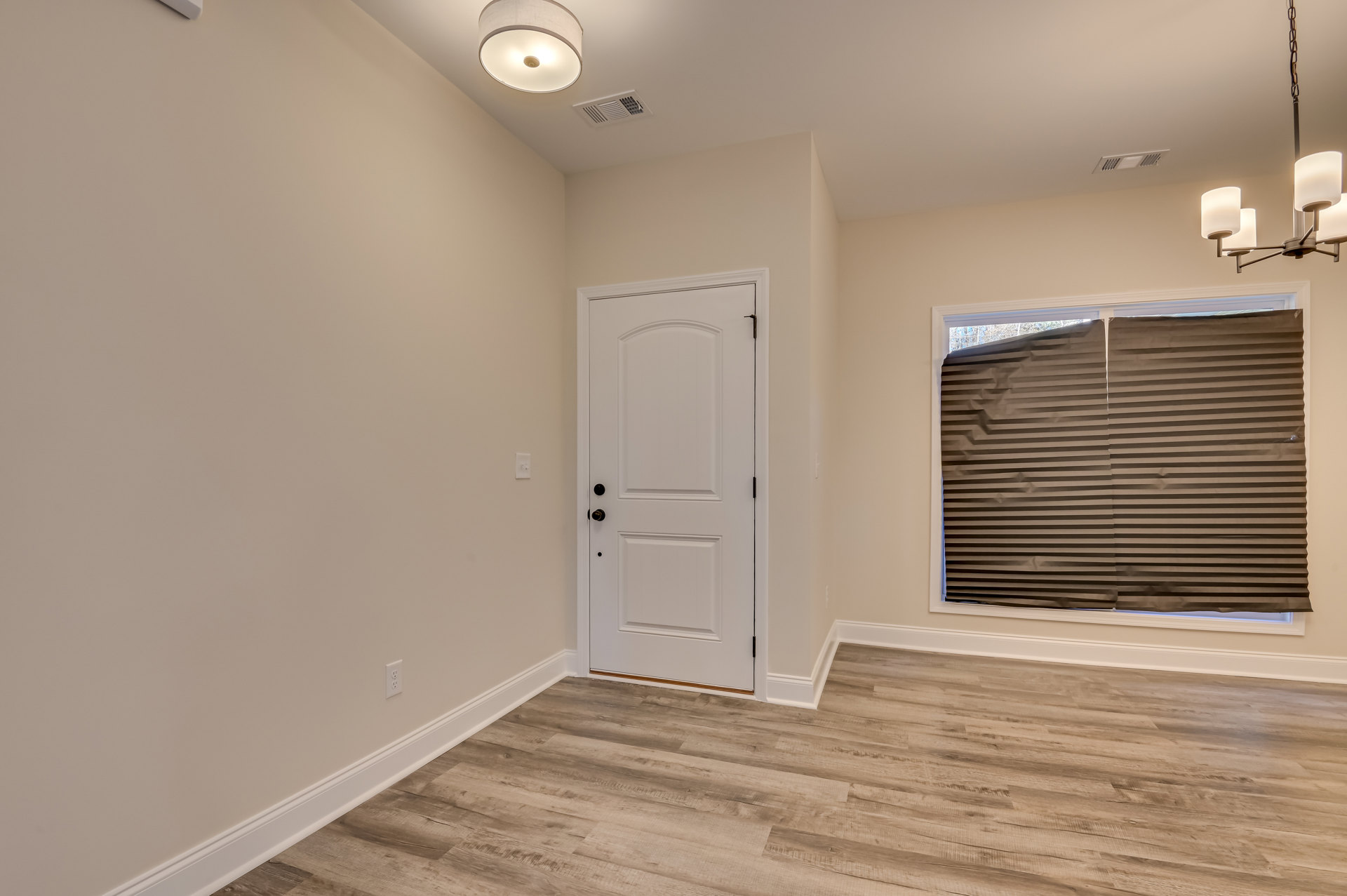 White paneled door with gold doorknob, brown window blind, wood laminate flooring, white ceiling, and wall-mounted light fixture with white shade