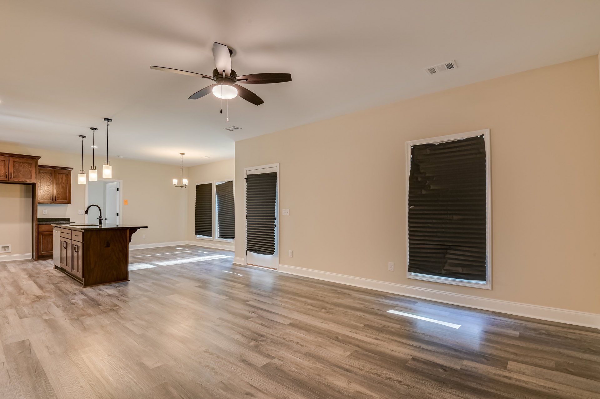 Open living area with wood flooring, ceiling fan with light, kitchen island featuring a sink, black blinds and brown shade on windows, neutral walls and plaster ceiling.