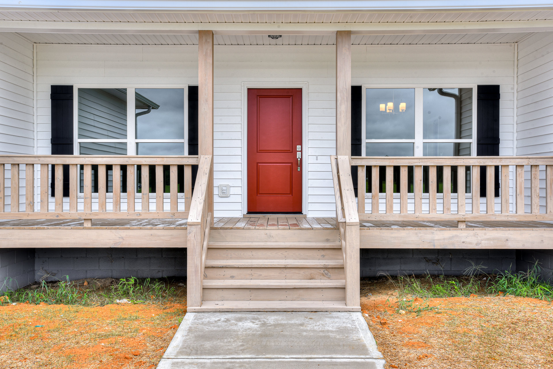 Front porch with wooden railing, red door featuring silver handle, concrete stairs and sidewalk, window with exterior light fixture, small plants along foundation