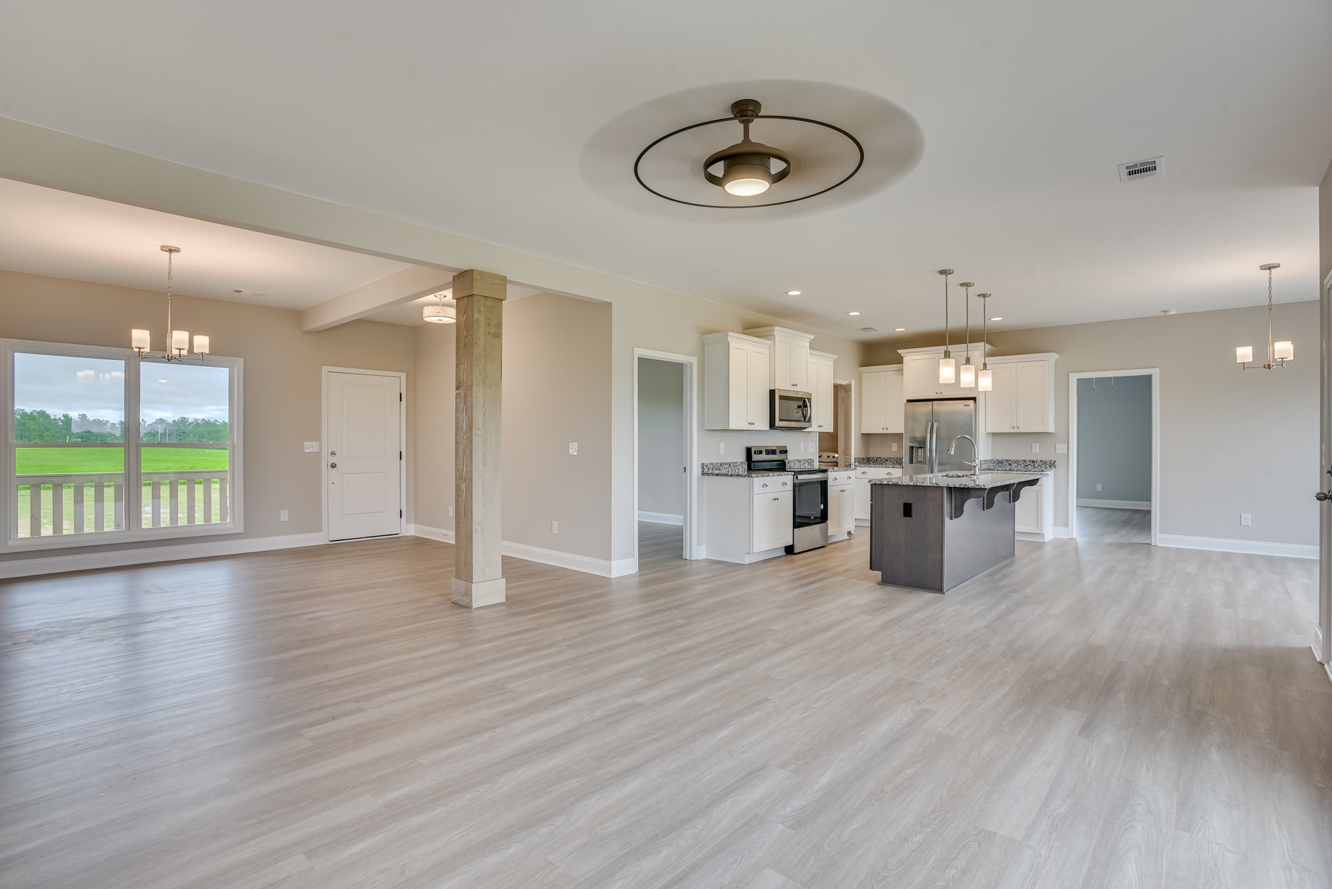 Spacious open-concept kitchen and living area featuring a central kitchen island, wood flooring, white door with silver handle, large window overlooking grassy field, and ceiling