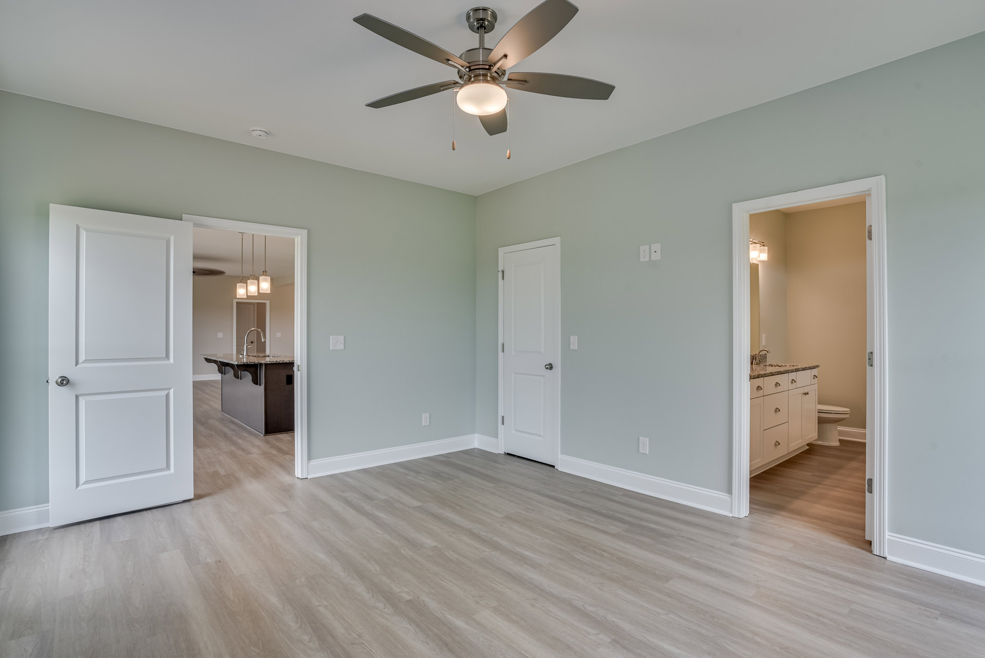 Bedroom with wood flooring, white walls, ceiling fan with light, white door with silver knob, white cabinet with silver handles, and a toilet visible in the corner