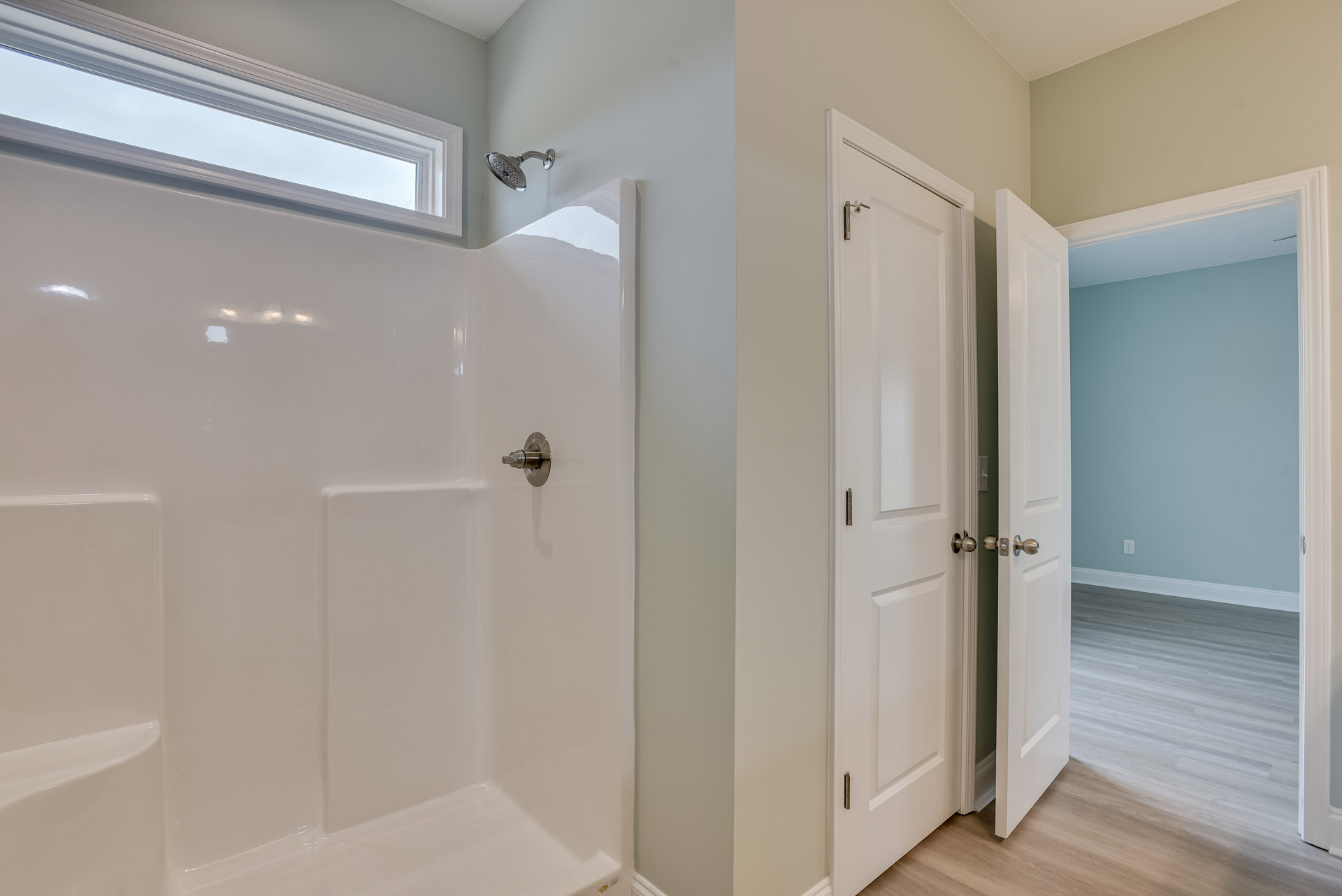 Modern bathroom featuring a glass-enclosed shower with chrome showerhead, white paneled door with brushed metal handle, light gray tile walls, and a frosted window allowing natural
