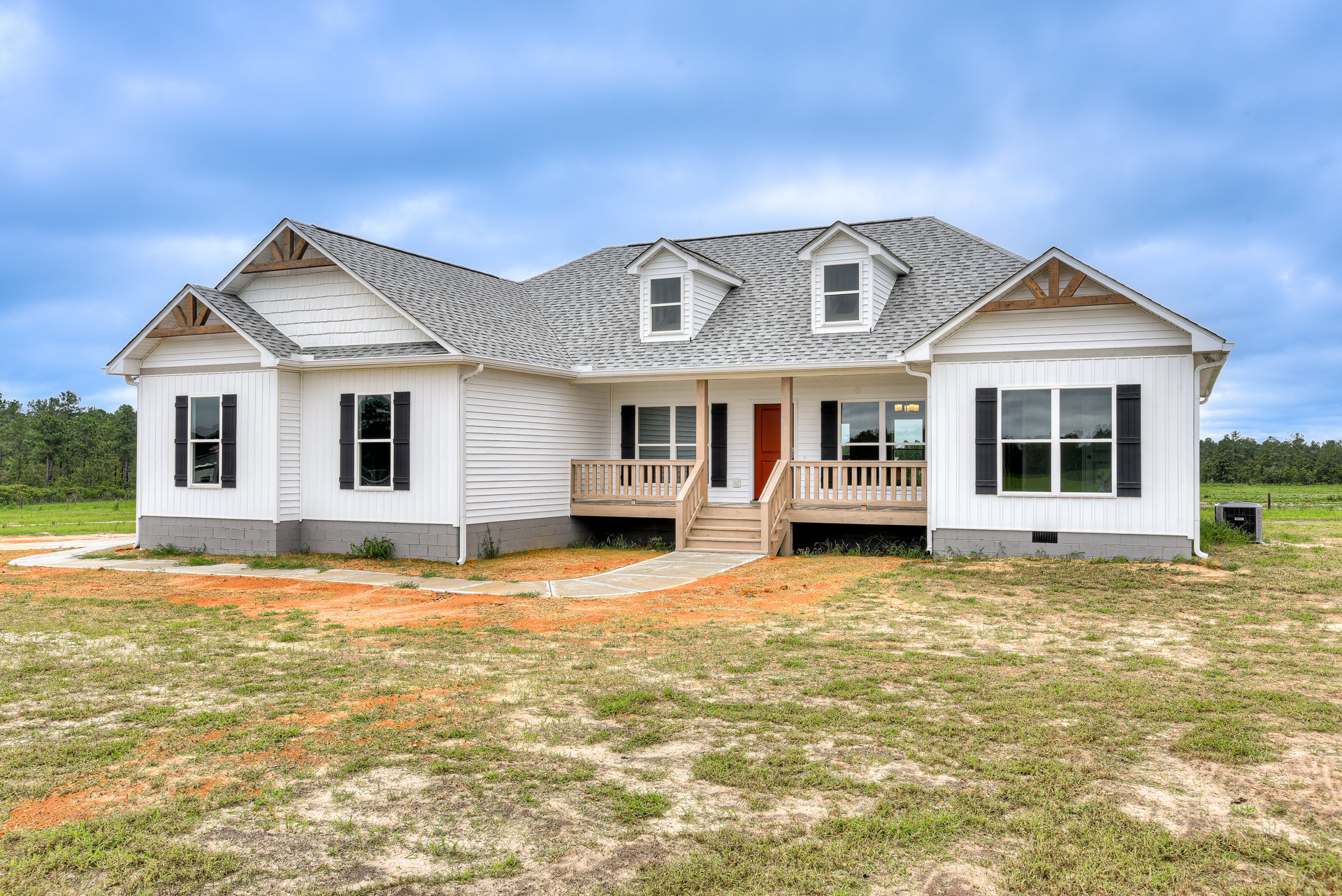 White siding house with red front door, covered porch, wooden bench, grassy yard, walkway, large windows reflecting trees, cloudy sky overhead