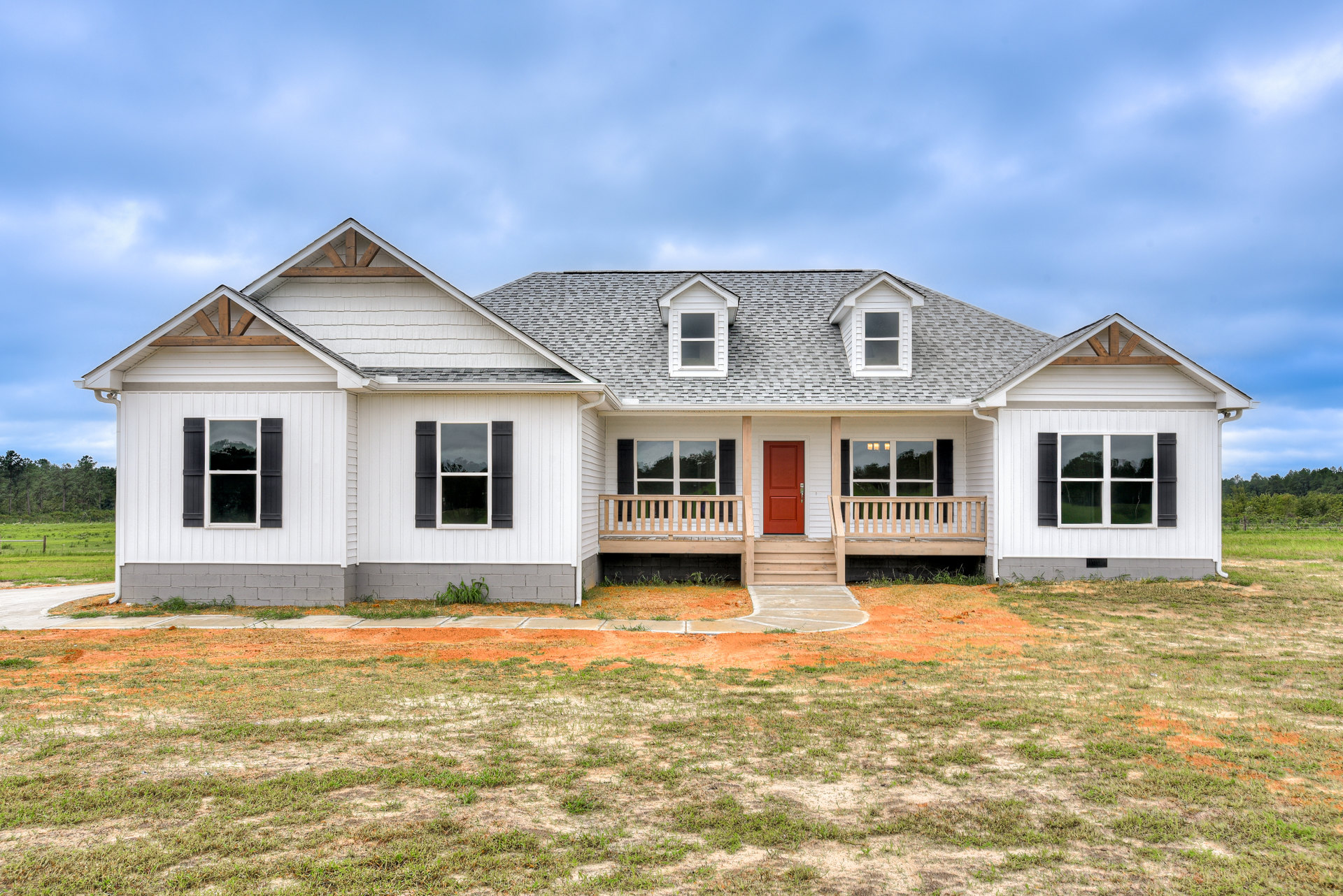 White siding house with black shuttered windows, red front door, wooden bench on covered porch, green grass yard under partly cloudy sky