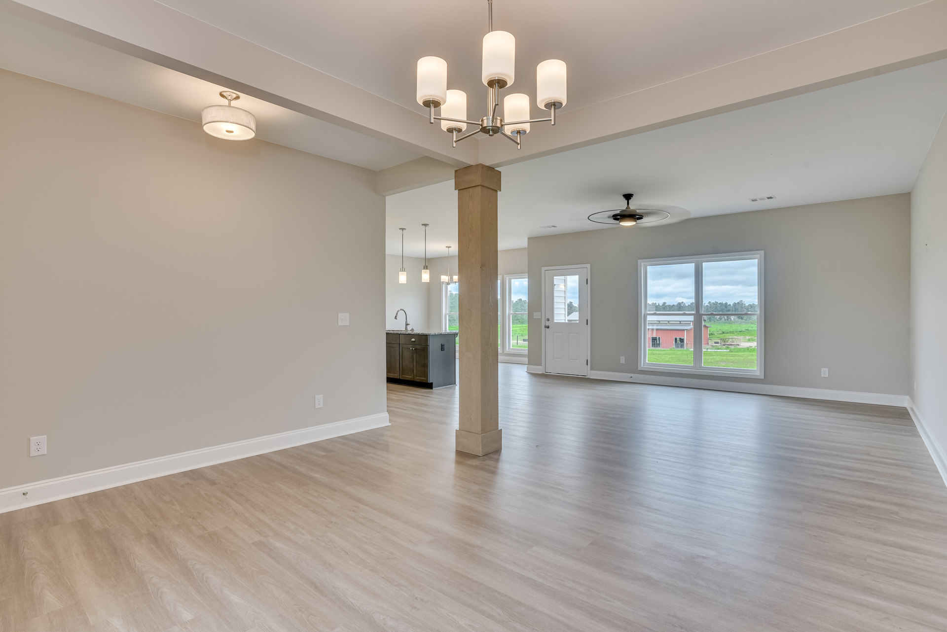 Spacious room featuring a prominent white pillar, wide plank wood flooring, white door with glass window, large window overlooking a farm and barn, plaster walls, ceiling lamp, and