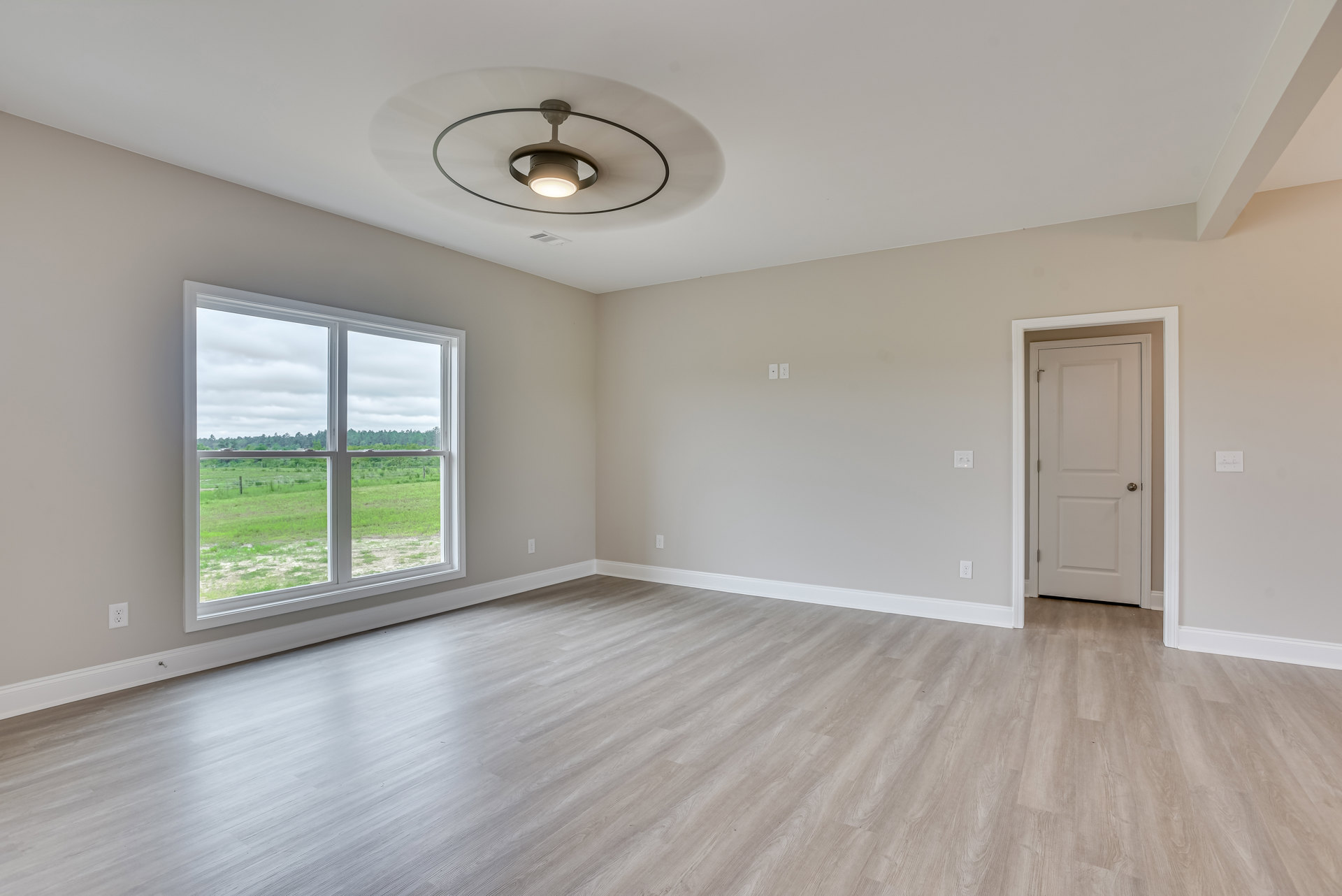 Sunlit room with wide window overlooking grassy field, wood plank flooring, white plaster walls, ceiling fan with light, and white door featuring silver knob