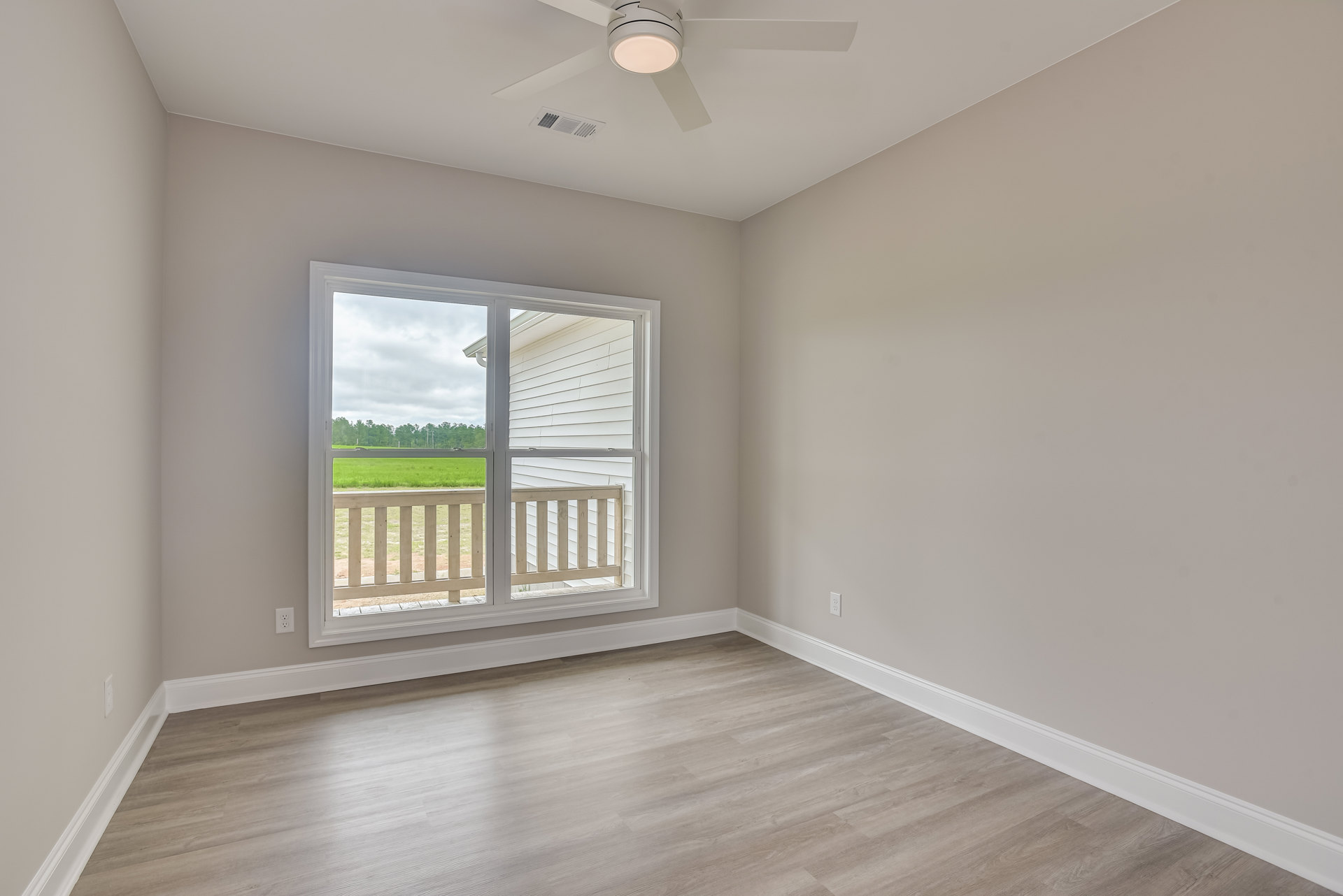 Bright room with wood flooring, white walls, large window overlooking a wooden fence and grassy yard, ceiling fan with light fixture mounted on smooth ceiling