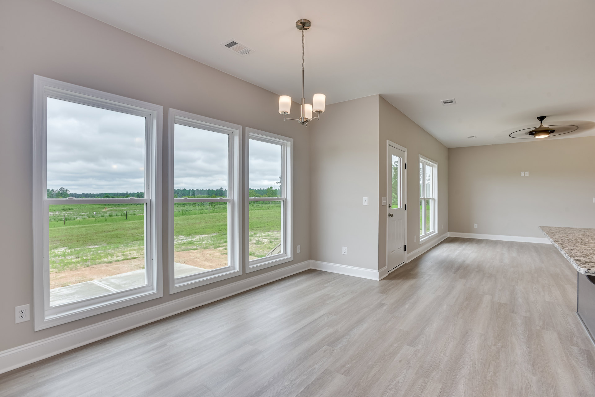 Bright room with large windows overlooking grassy field and trees, wood laminate flooring, white plaster walls, ceiling fan with light fixture