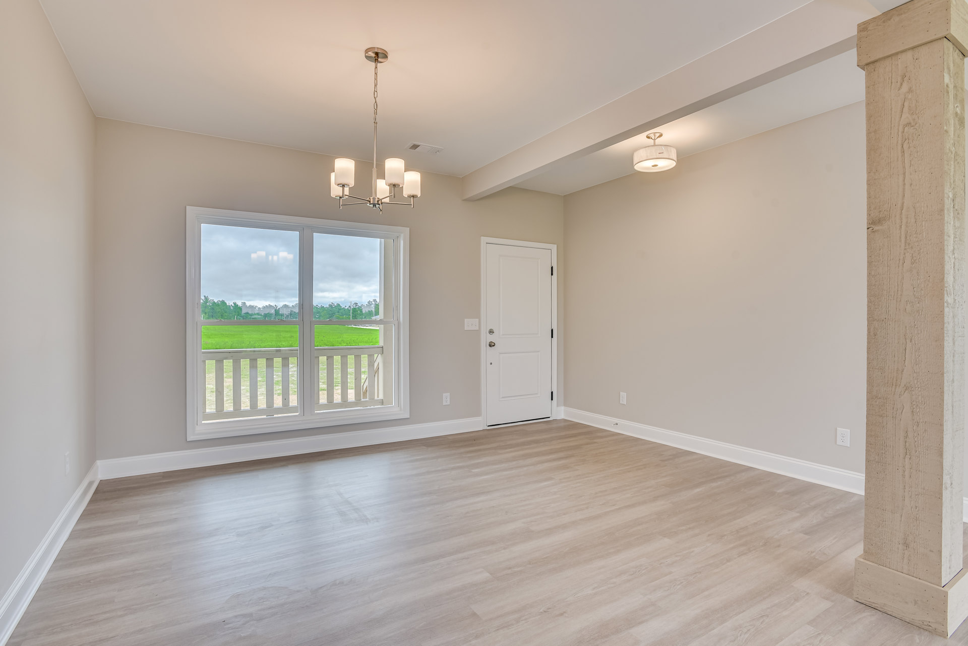 Wood flooring with white baseboards, white door featuring a silver handle, large window overlooking a grassy field, neutral plaster walls, and a modern lamp in the corner.