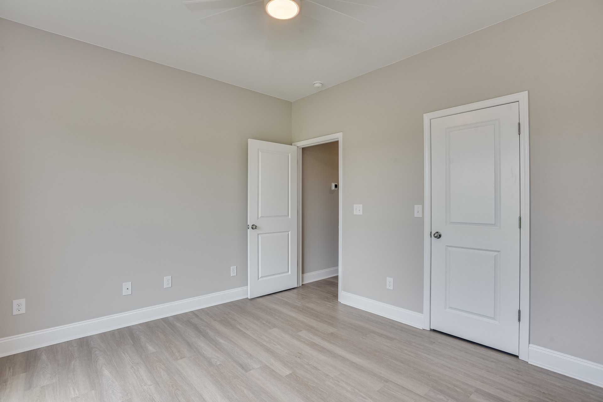 Room with wood flooring, white trim, two white doors with silver knobs, ceiling fan with light, and neutral plaster walls.