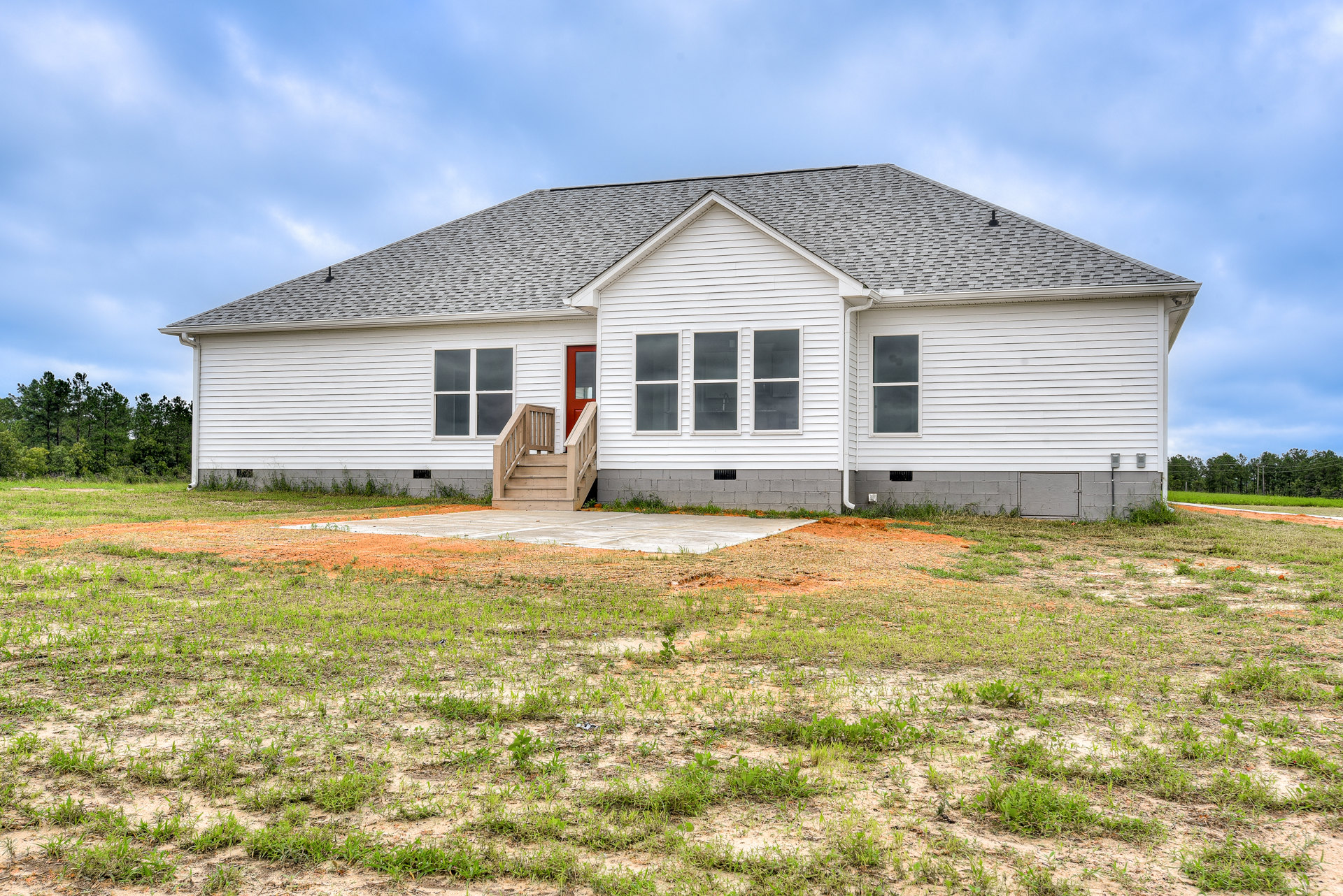 White farmhouse with red front door, three windows, wooden exterior stairs, surrounded by green lawn under partly cloudy sky