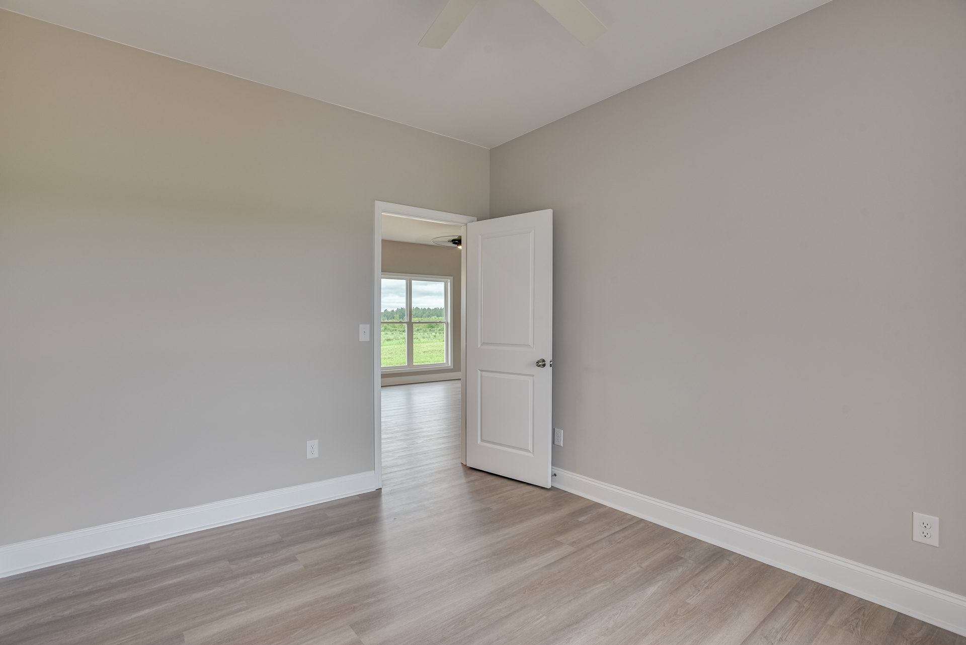 White room with laminate wood flooring, open white door with silver knob, ceiling fan, electrical outlet on plaster wall, window showing green trees outside
