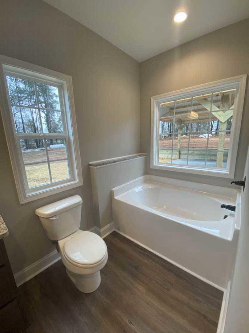Bathroom with white tile floor, freestanding tub, white toilet with lid up, large window overlooking green trees, neutral wall finishes