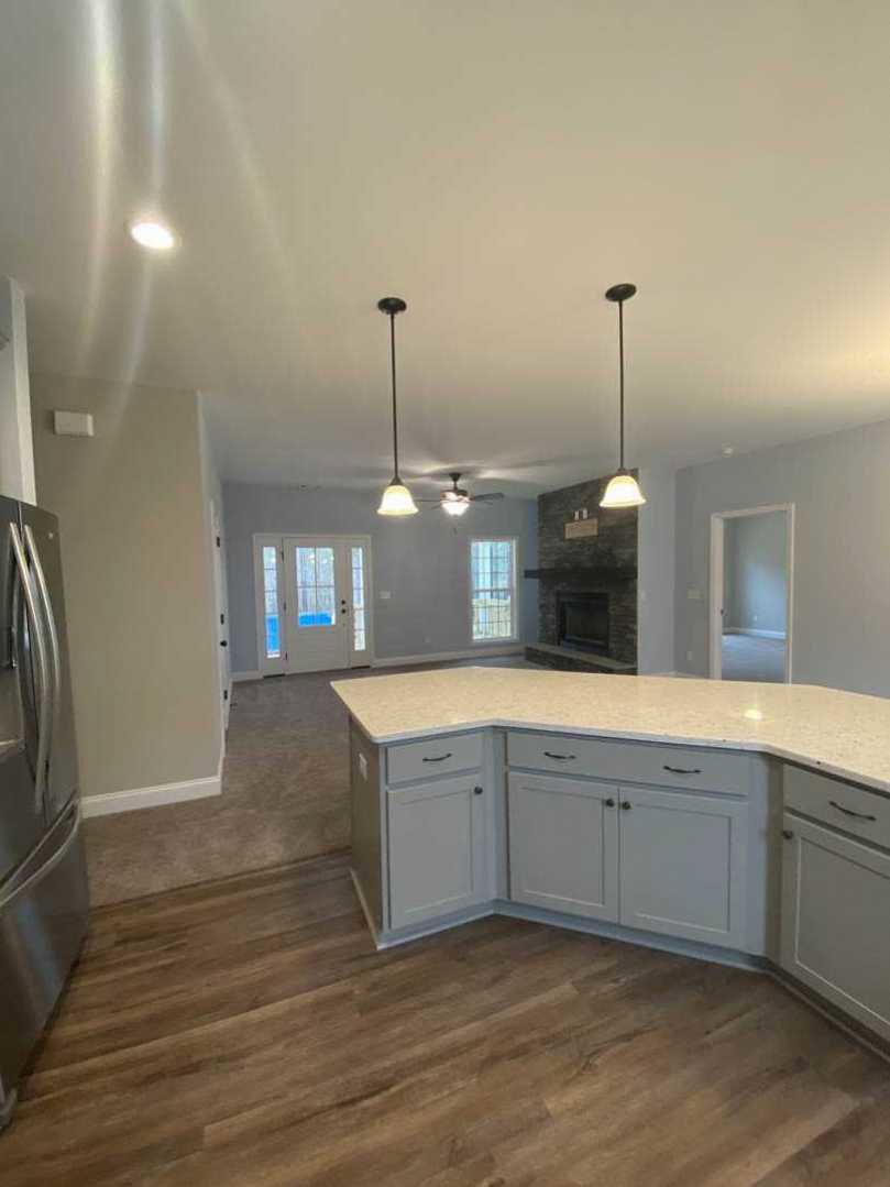 Open kitchen with stone fireplace, oversized island with marble countertop, wood flooring, white cabinetry, glass-paneled door, ceiling recessed lights, and neutral curtains