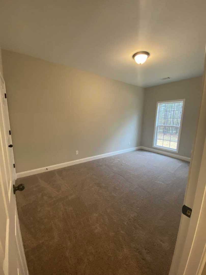 Carpeted room with white-framed window, white door featuring black hinge and silver knob, ceiling-mounted light fixture, smooth plaster walls