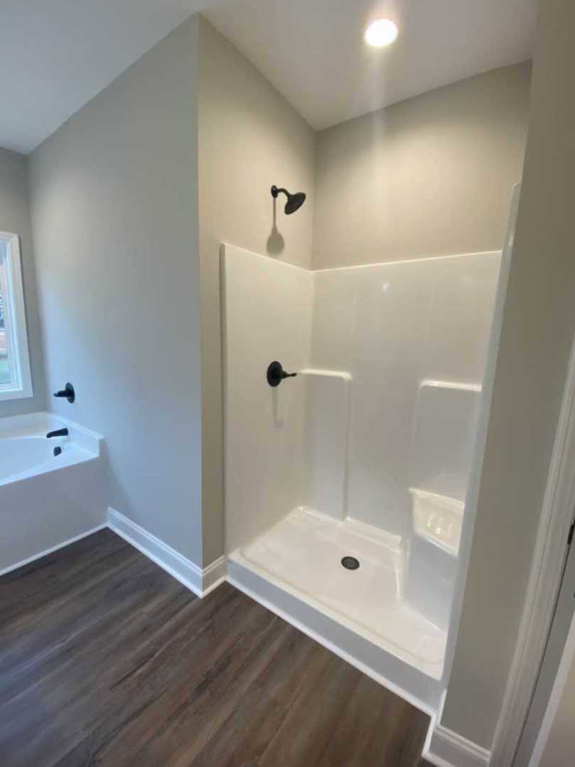 Bathroom with freestanding white bathtub, glass-enclosed shower featuring chrome showerhead, light wood flooring, white baseboards, and neutral tile walls