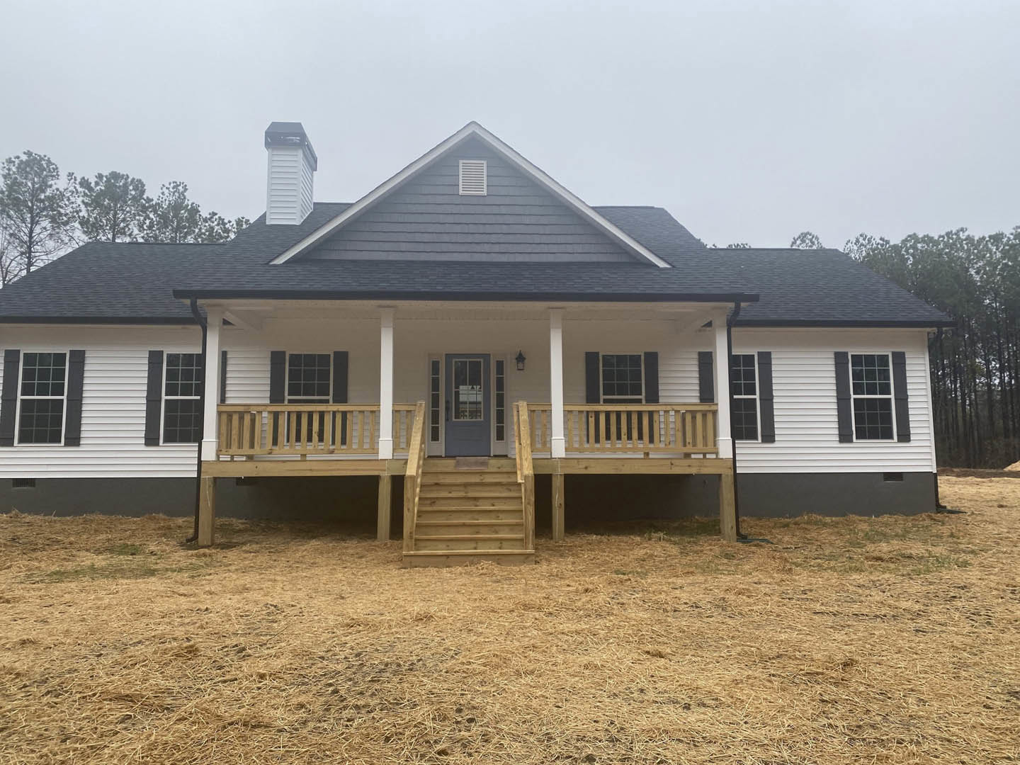 White siding house with black roof, grey door, covered porch, and wooden stairs leading to a wood deck; windows and trees visible in background.