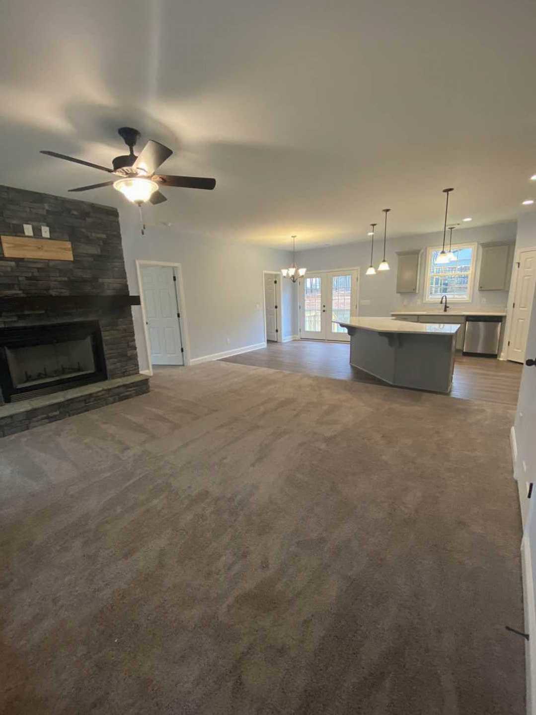 Open concept living room with carpet flooring, white fireplace featuring a glass window, ceiling fan with light, adjacent kitchen with visible countertop, white door framed in