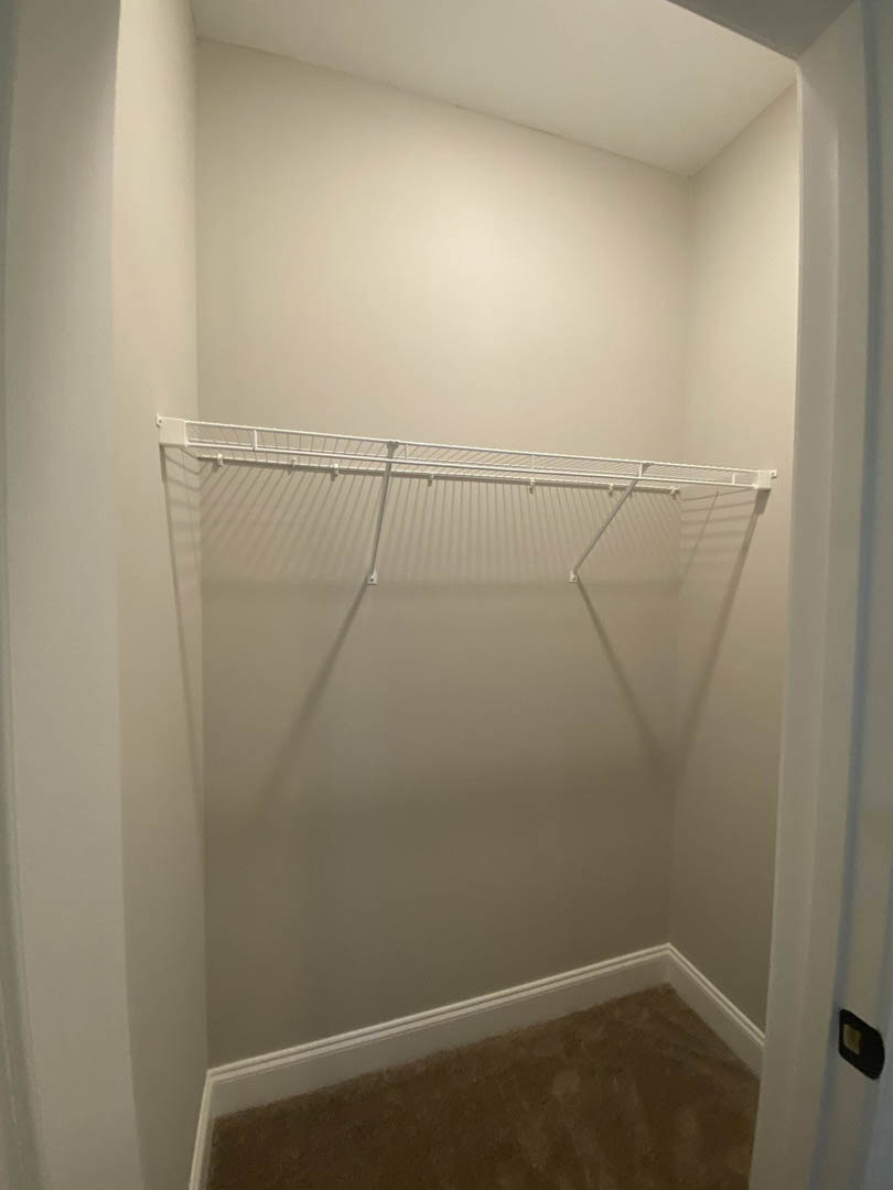 White closet with built-in shelf, white metal hanging rod, black rectangular object with gold label on wall, white plaster walls and ceiling.