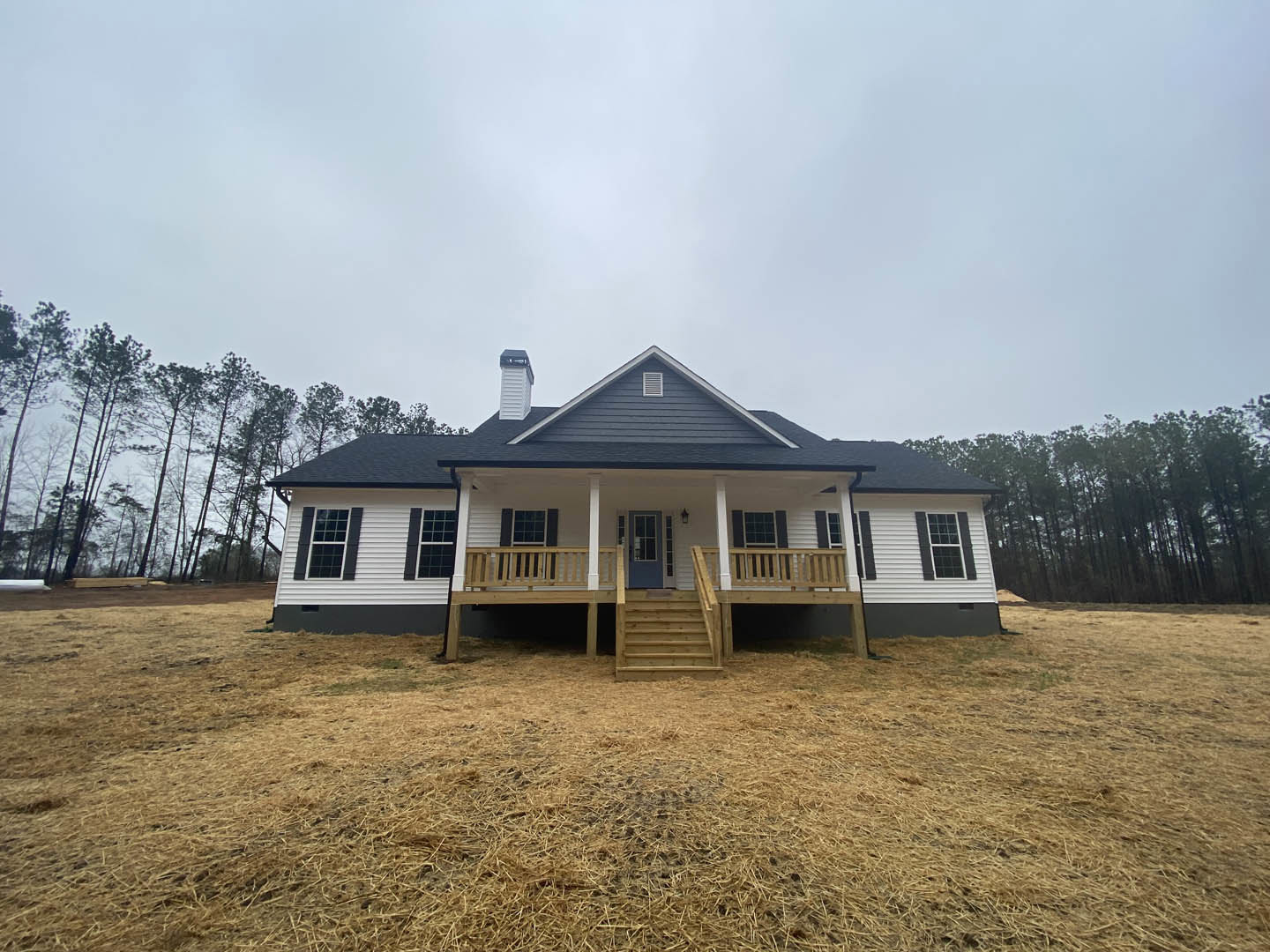 Two-story house with covered front porch, wooden steps, tall trees in background, vented roof, and windows facing a partly cloudy sky