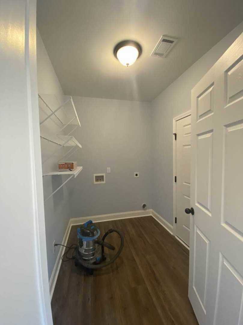 Vacuum cleaner on light wood laminate floor near white wall and door with black handle, ceiling light fixture overhead, white vented object against wall