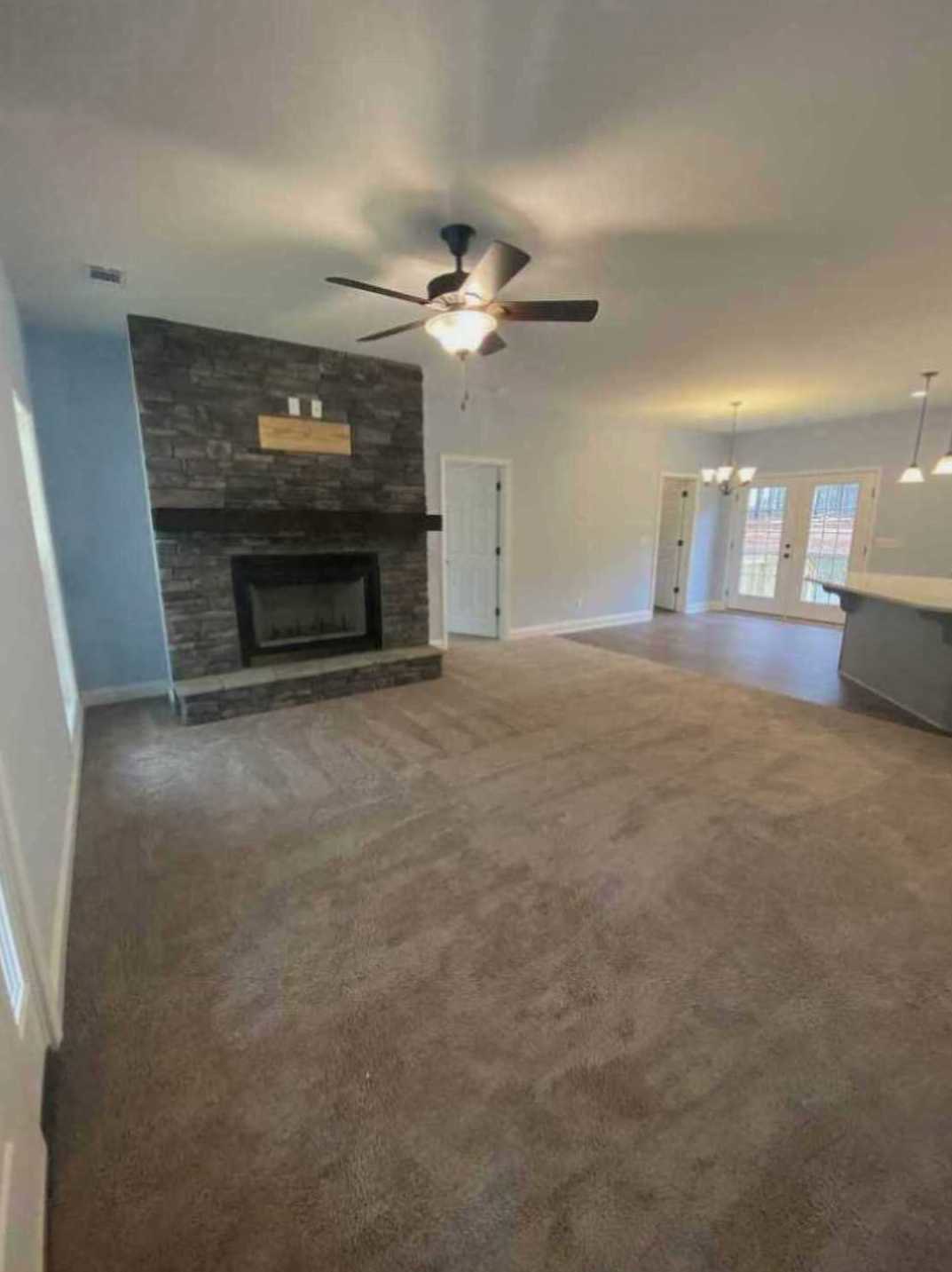 Carpeted living room featuring a central fireplace with white mantel, ceiling fan with light fixture, white double doors with black handles, plaster walls, and neutral finishes.