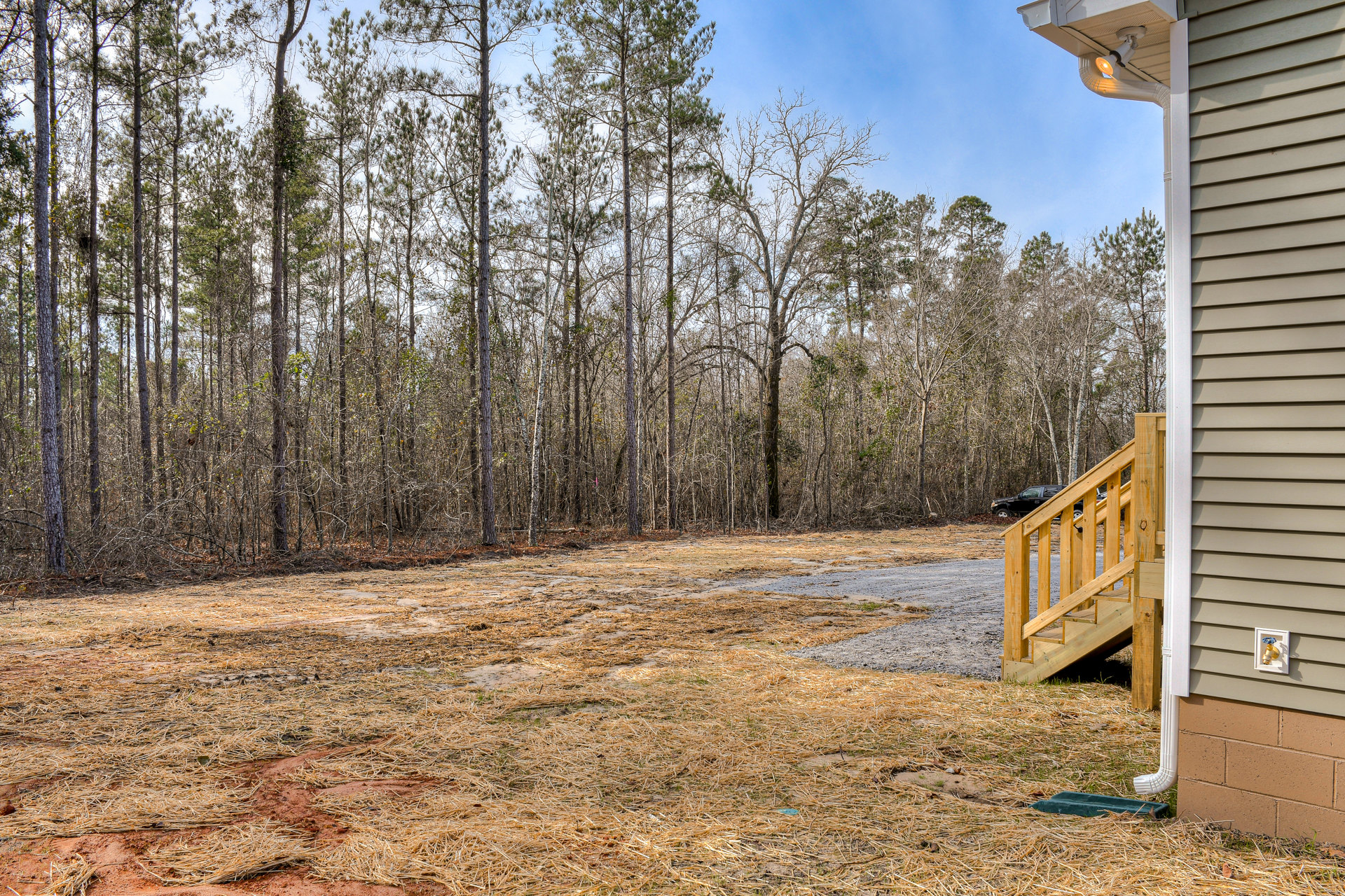 Wooden staircase with black metal railing leading to front entrance of house, surrounded by dry grass, trees, and wood fencing, under partly cloudy sky.
