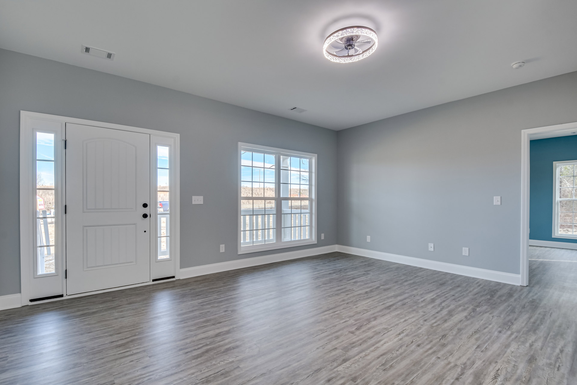 Wood floor room with white-framed window overlooking a field and blue sky, ceiling fan with circular light, white walls, and single door.