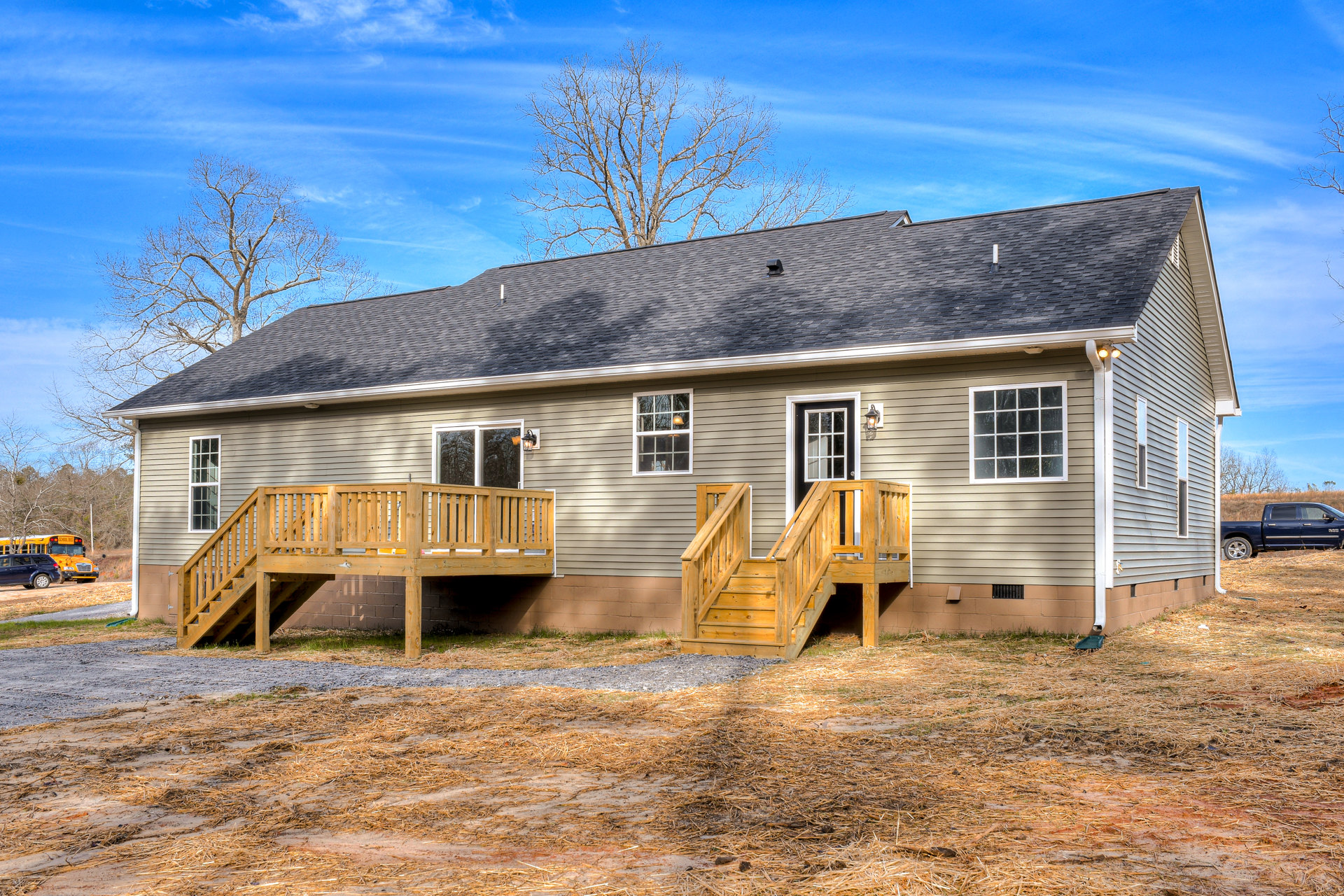 Wooden deck with exterior stairs leading to entry, multi-pane windows, warm interior lighting, blue pickup truck parked on dirt driveway, surrounded by trees and plants under