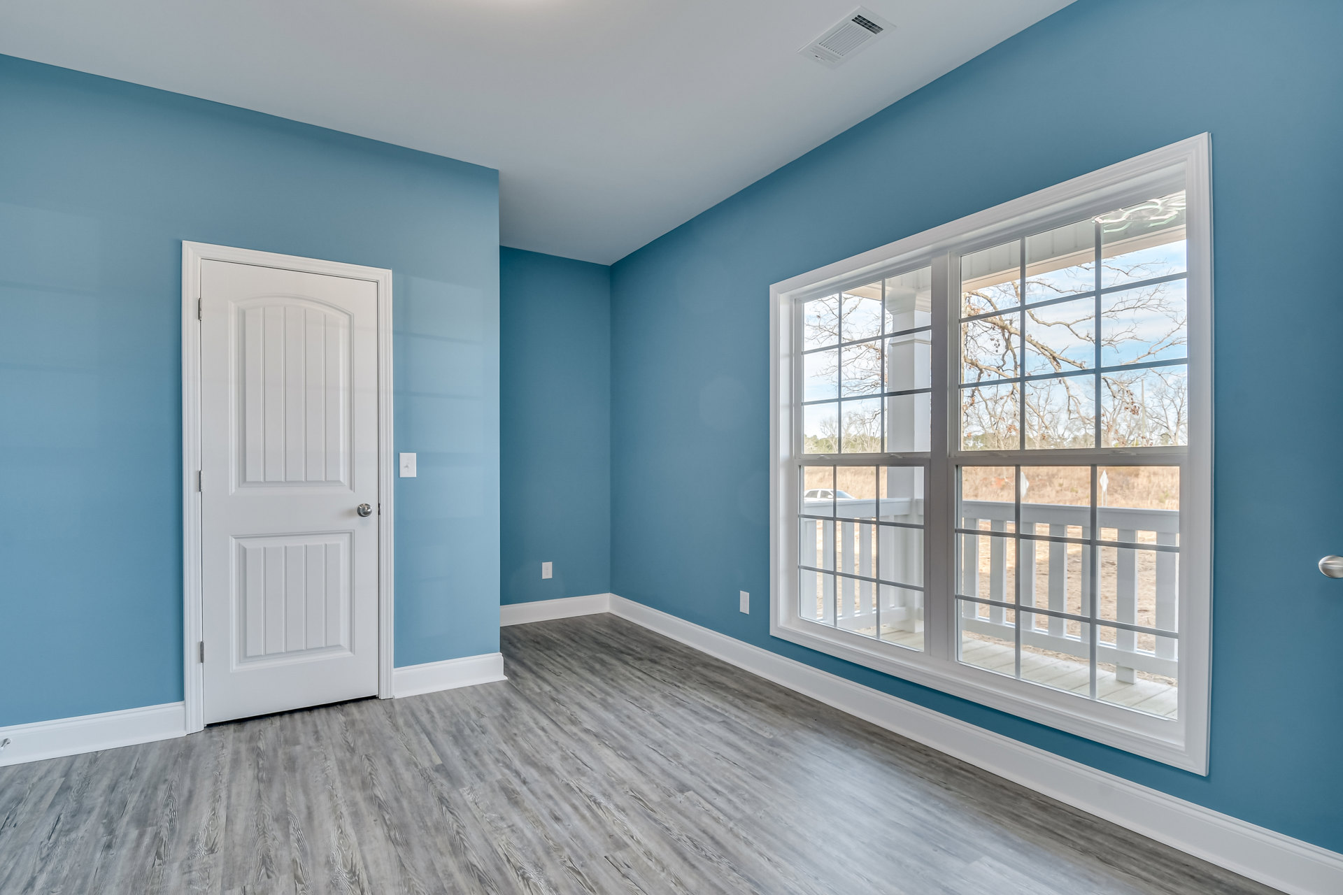 White paneled door with silver knob, large window overlooking porch and parked car, light wood flooring, smooth white walls