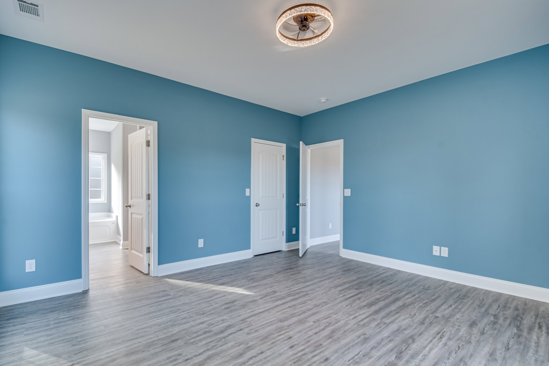 Blue-painted walls, white paneled doors with silver handles, wood laminate flooring, ceiling fan with light fixture, white molding, adjacent bathroom with white bathtub and door
