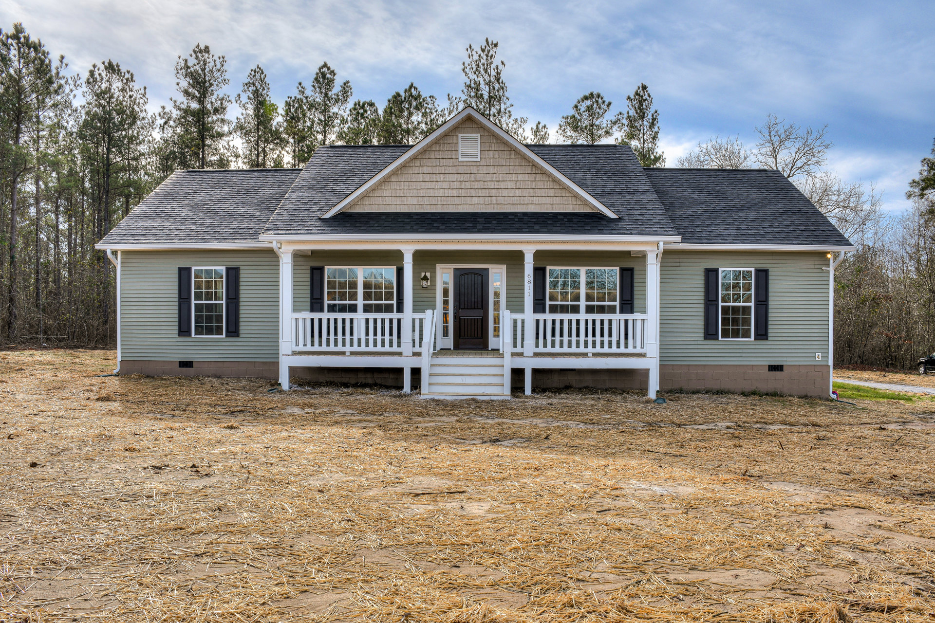 White cottage-style home with covered front porch, black entry door, white-framed windows reflecting nearby trees, and cloudy sky overhead