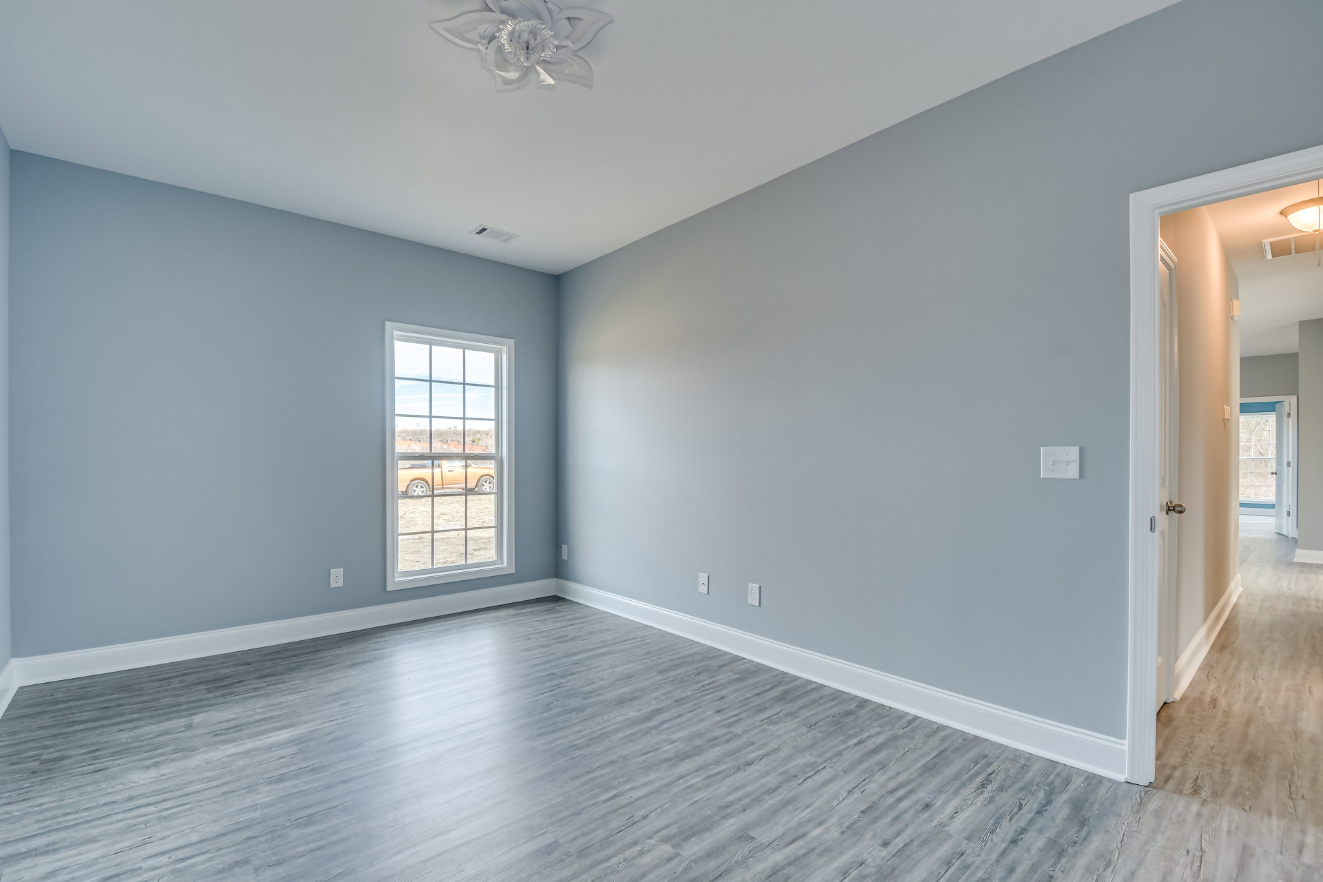 Bright room with grey wood flooring, white baseboards, large window overlooking parked truck, light switch on wall, ceiling fan with light fixture