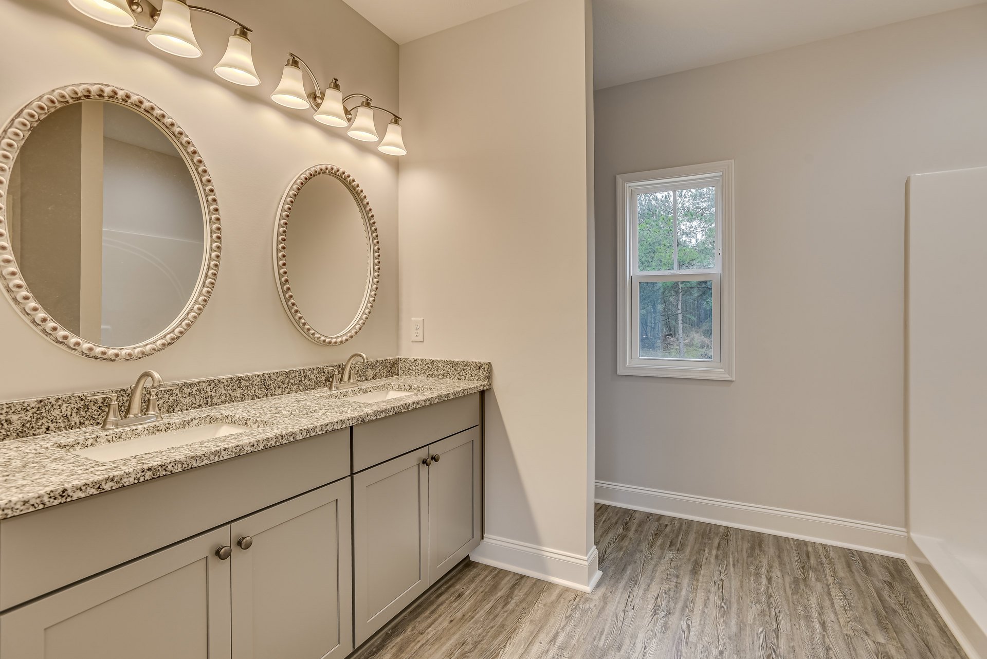 Marble bathroom countertop with two sinks, white framed mirrors, oval mirror accented with brown and white beads, chrome faucets, white cabinetry, window overlooking trees, and