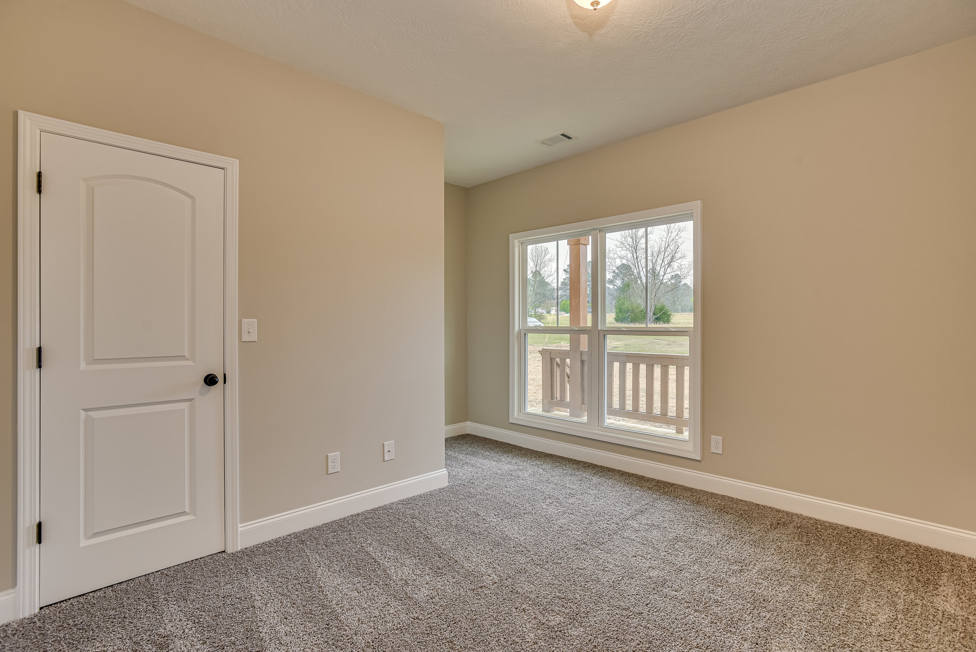 White paneled door with black handle, beige carpeted floor, large window overlooking leafless trees and grassy yard, white walls with crown molding