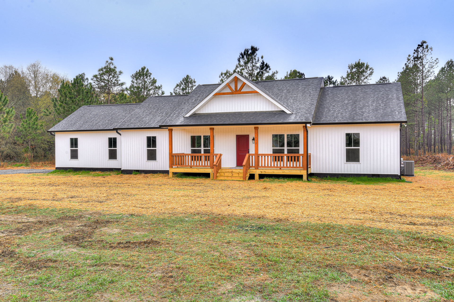White farmhouse with red front door, wooden porch and deck, expansive green lawn, mature trees, and large windows under a blue sky