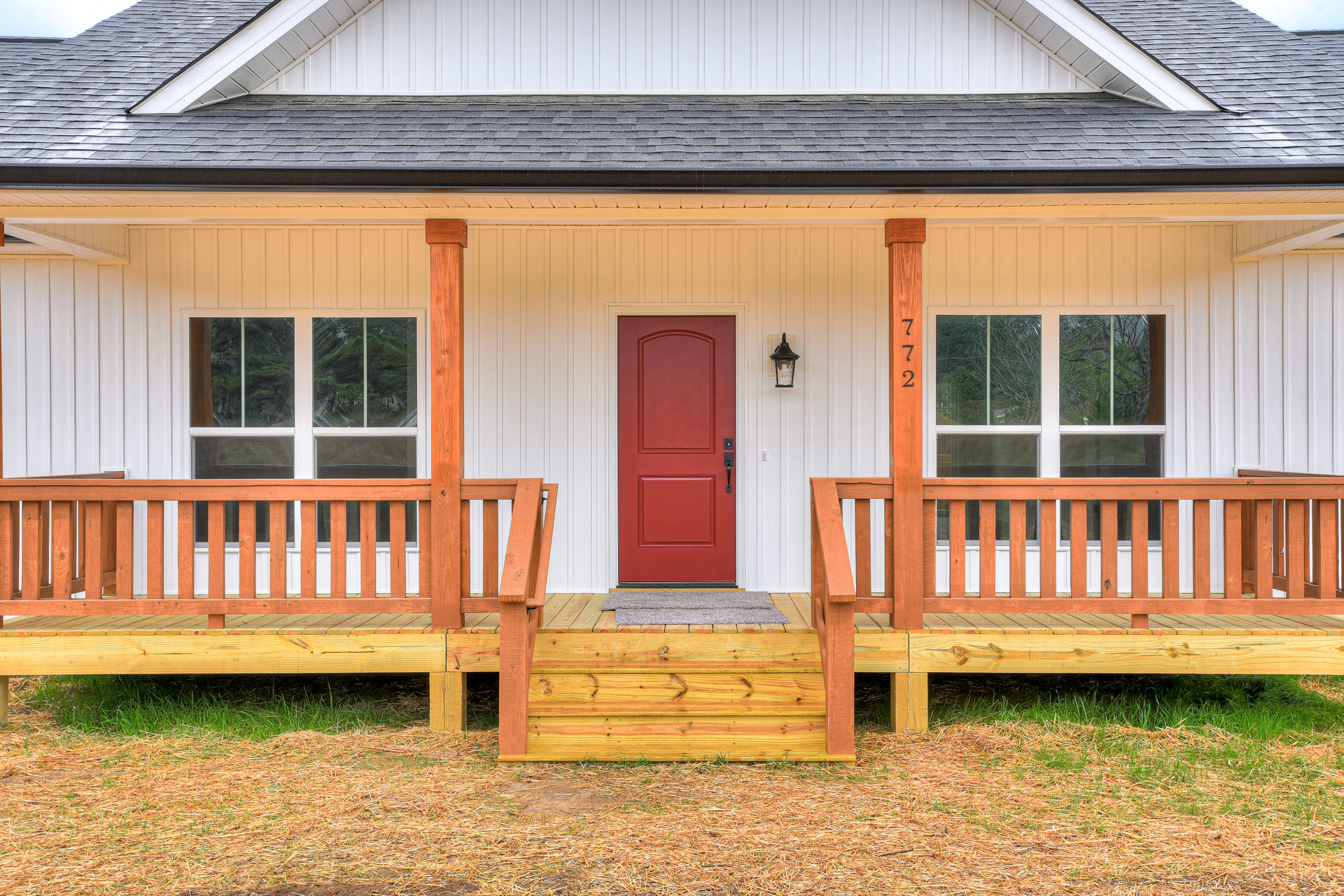 Red front door with black handle, wooden porch and steps, wood siding exterior, railing, green grass lawn