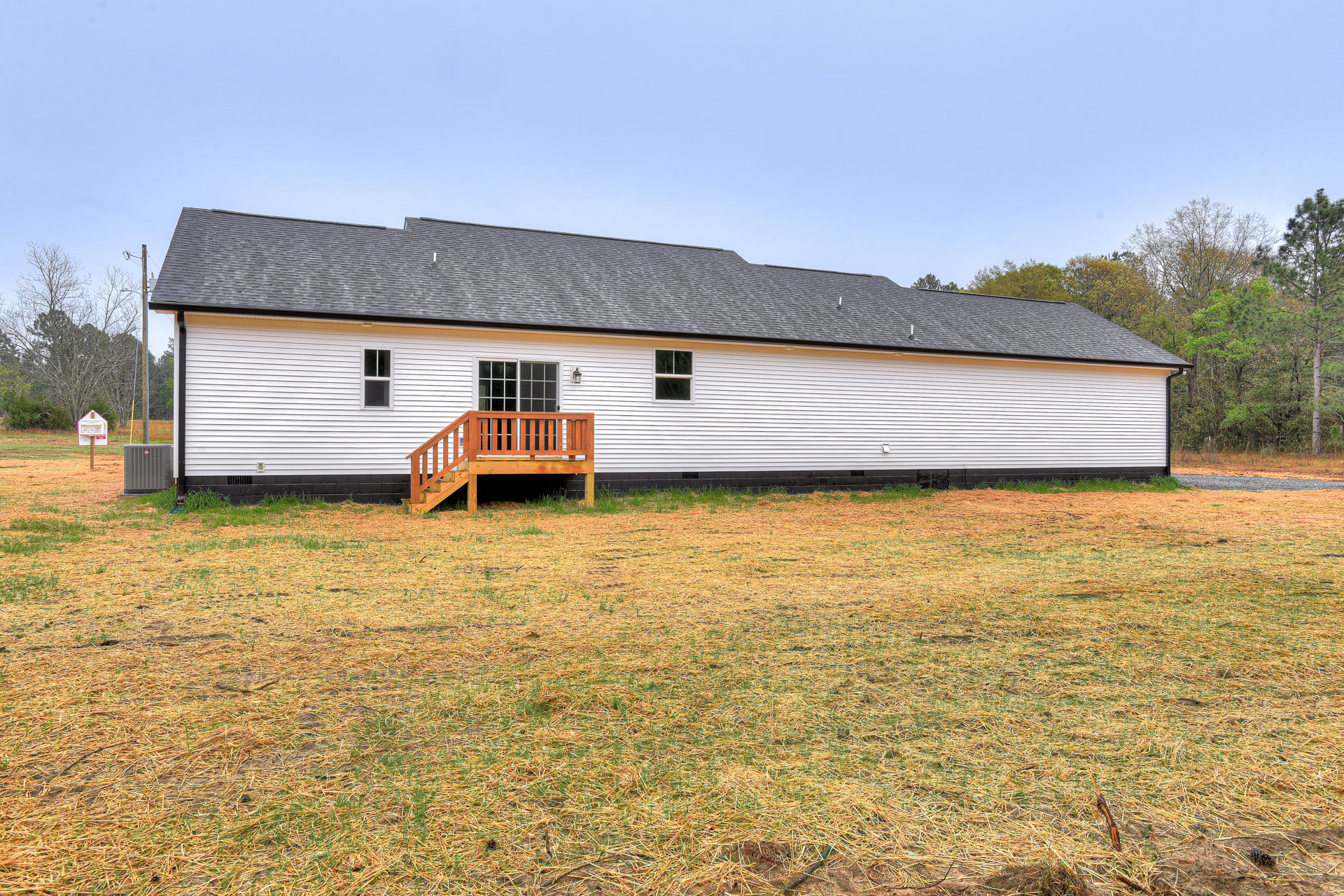 White siding house with large wooden deck, stairs leading to grassy lawn, windows visible, surrounded by open green yard and trees.
