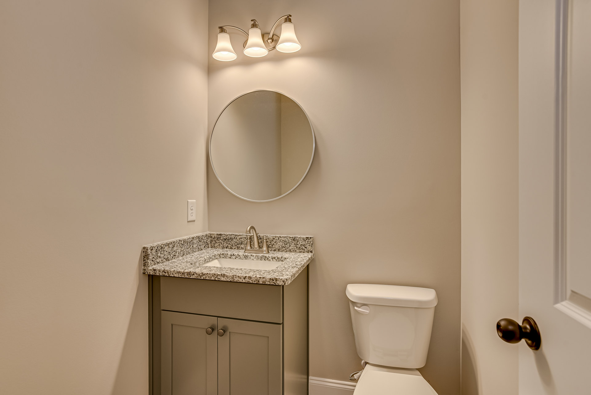 White bathroom with tile walls, round mirror above a rectangular sink, modern light fixture, white toilet with closed lid, dark cabinet doors, and chrome door knob