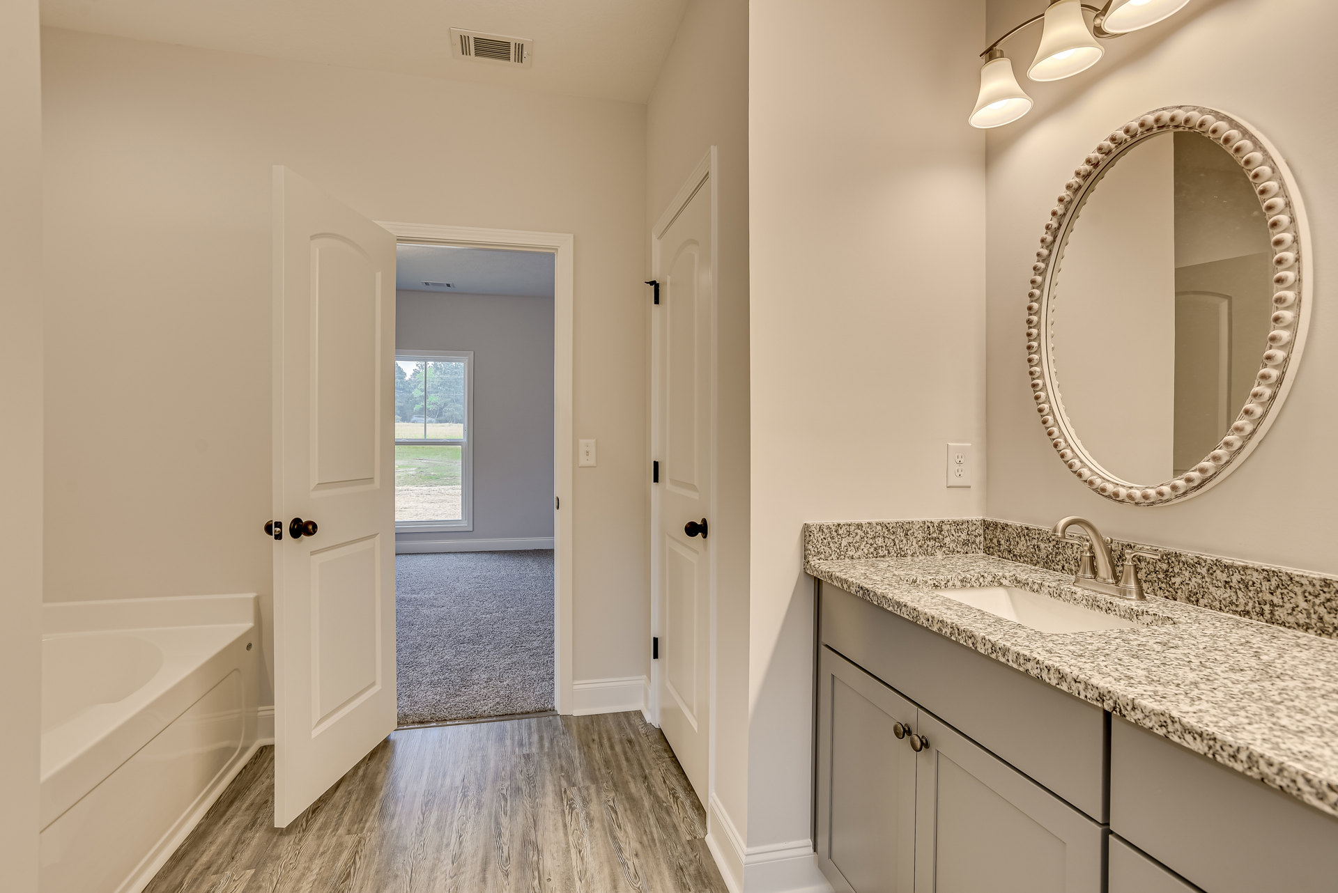 Bathroom with marble countertop sink, large mirror, white freestanding bathtub, wood flooring, and window overlooking grassy field.