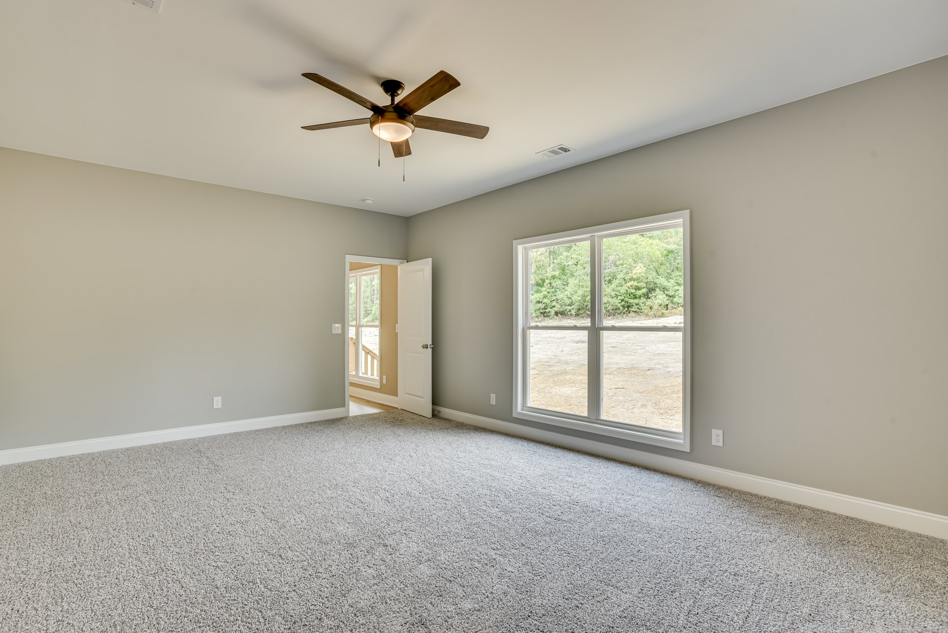 Carpeted room with white walls, ceiling fan with light fixture, large windows showing leafy trees, open white door with silver knob, crown molding along ceiling.