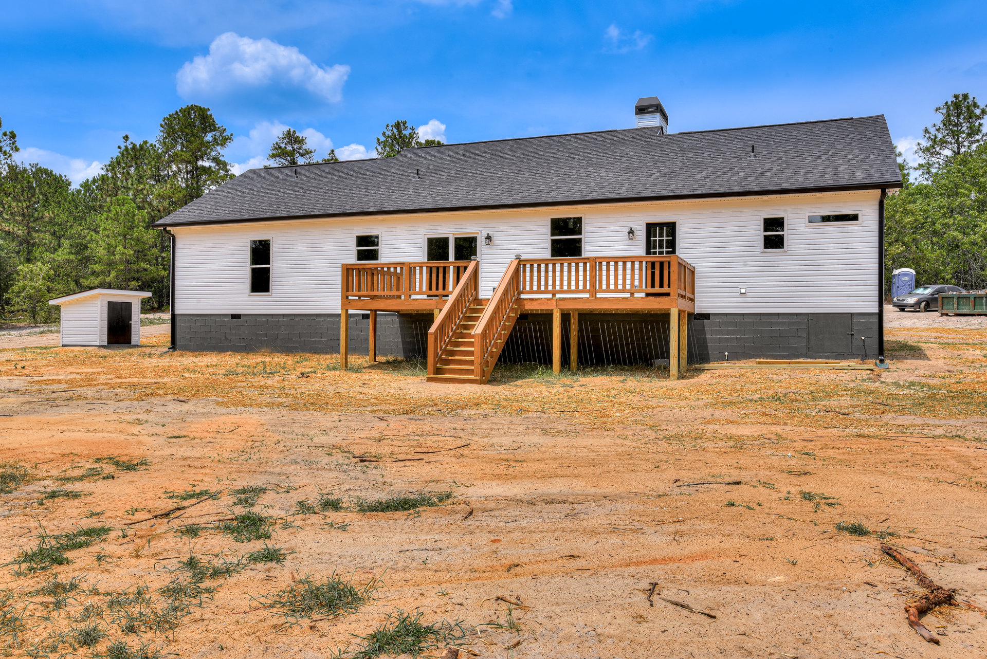 White house with black door, wooden porch and stairs, surrounded by dirt and grass, trees in background under cloudy sky