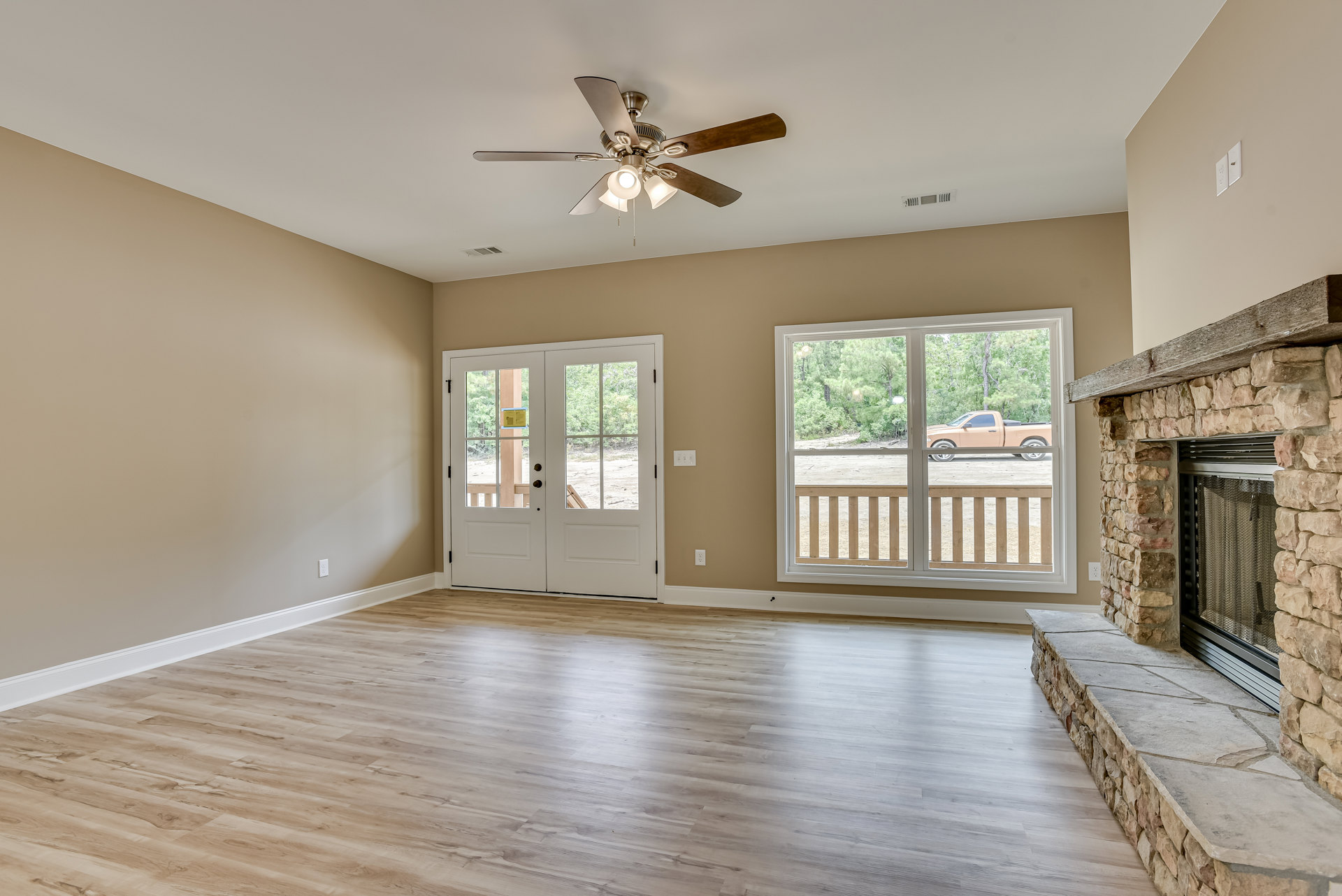 Living room with wood flooring, stone fireplace, ceiling fan with lights, glass-paneled double doors, and window showing parked truck outside.