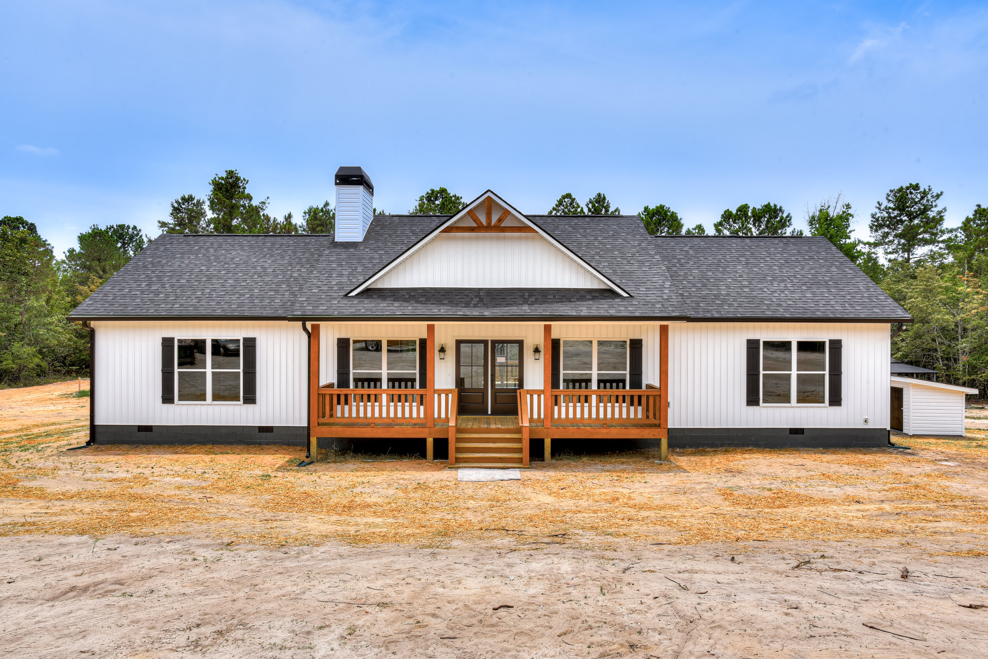 Wooden porch with white railing and trim, wooden steps leading to entry, cottage-style house set along a dirt road, trees and cloudy sky in background