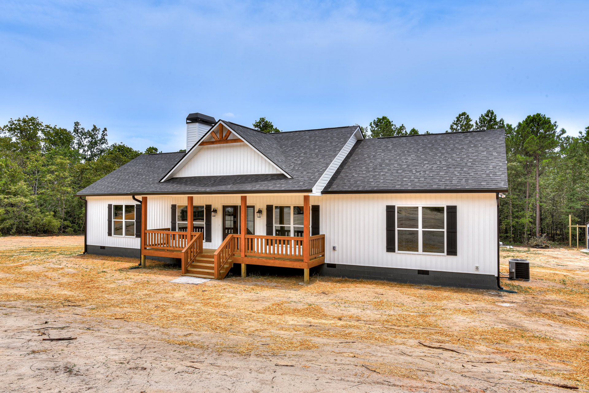 White siding house with covered porch, wood deck, railing, ground-mounted heat pump, large window reflecting dirt road, gabled roof, and surrounding trees.