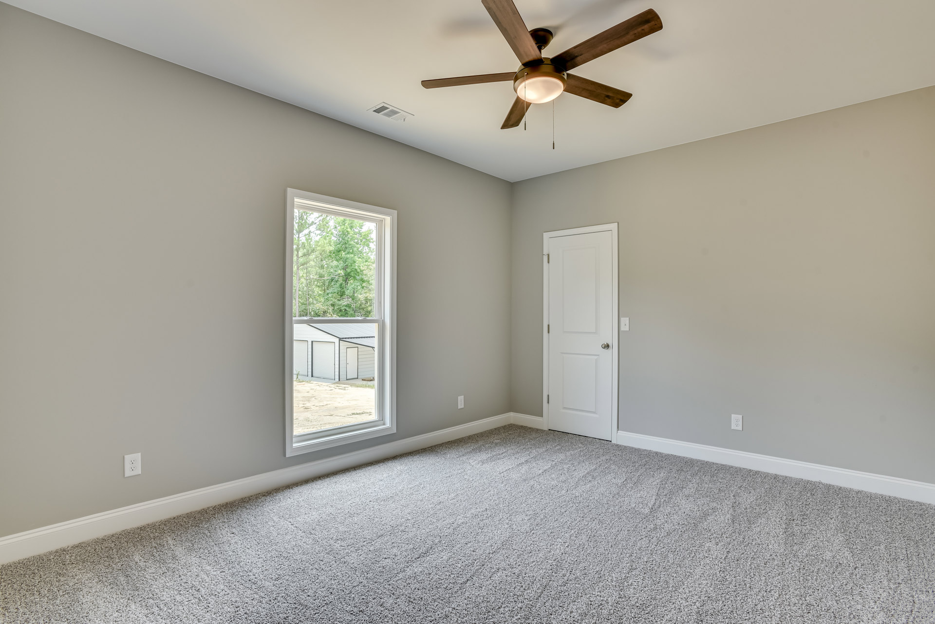 Carpeted room with white walls, ceiling fan with light fixture, large window showing trees and neighboring house, white door with silver knob, crown molding along ceiling.