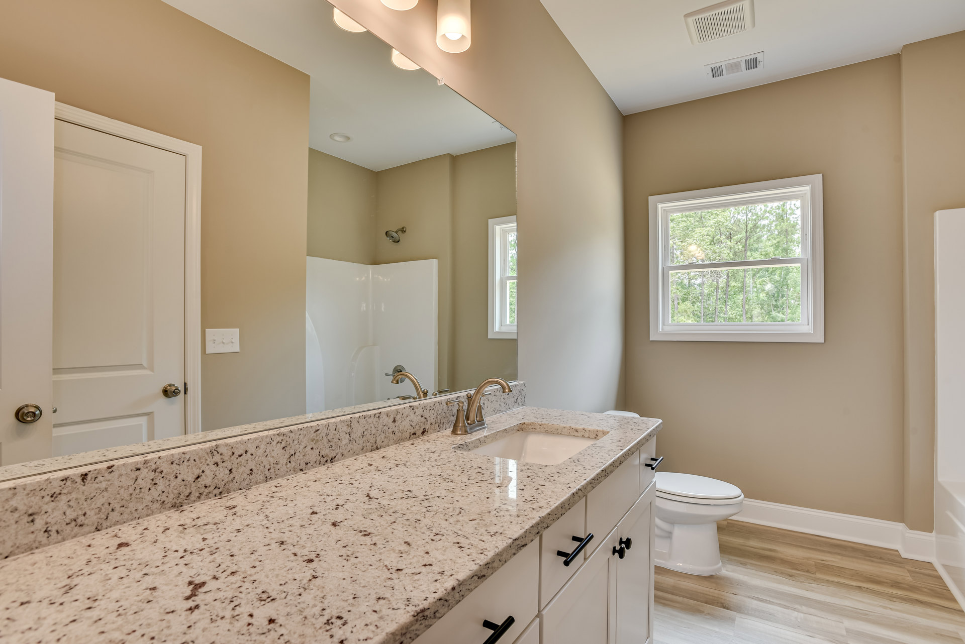 Bathroom with marble countertop, undermount sink, chrome faucet, white toilet with lid closed, white walls, large window overlooking trees, glass shower enclosure
