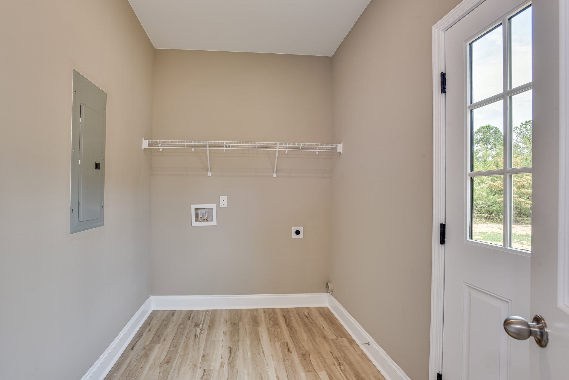 Wood floor with white baseboard, white door featuring black square inset, white-framed window showing trees outside, close-up of silver doorknob, two exposed pipes within white