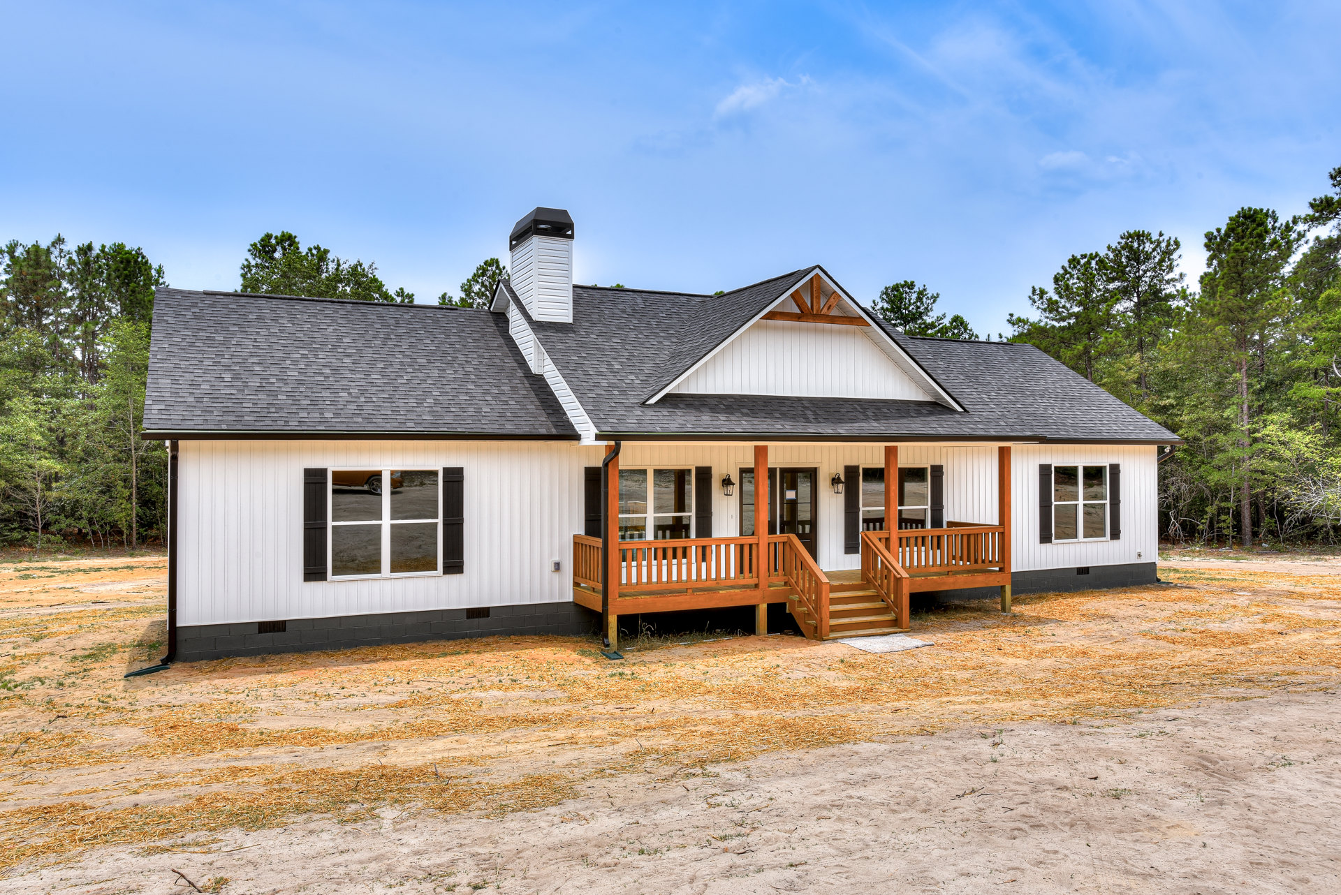 White cottage-style home with wood porch, wooden stairs, driveway, chimney, large windows reflecting sky, surrounded by trees and dirt landscaping